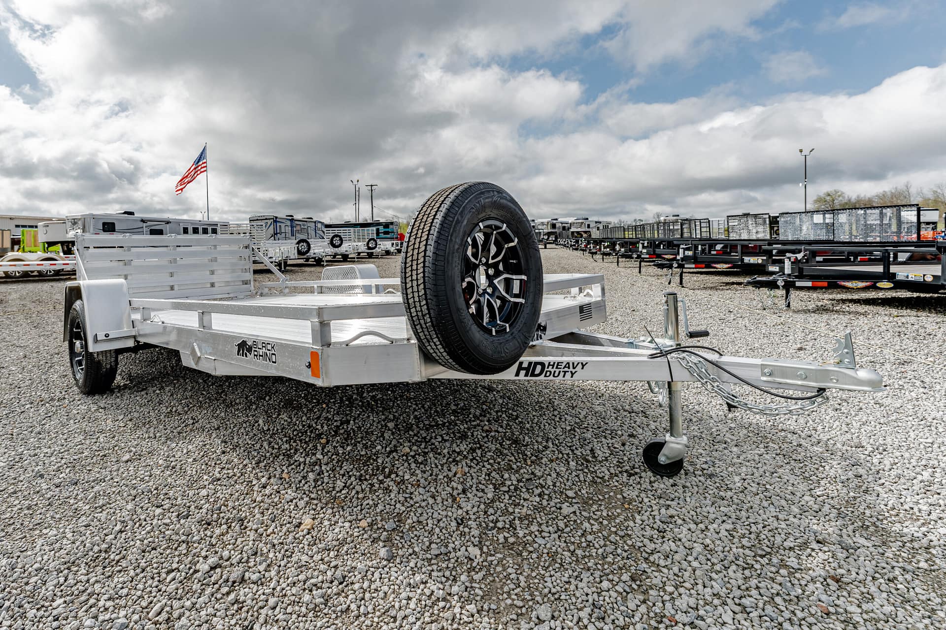 Aluminum utility trailer with a mounted spare tire in a gravel lot, showing a hitch and wheel, with an American flag and other trailers in the background