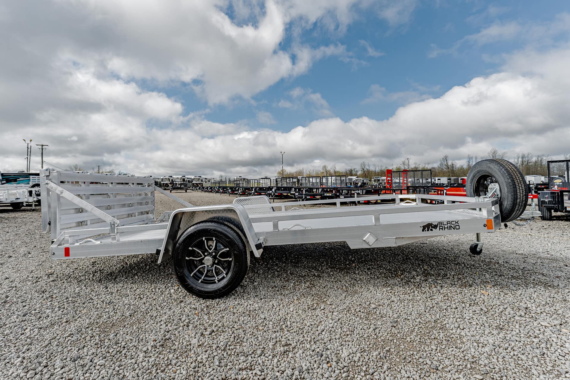 Aluminum flatbed trailer with spare tire on right, brand 'Black Rhino', parked on gravel lot with other trailers in background