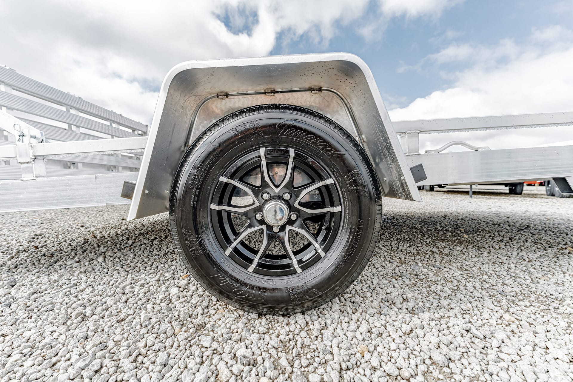 Front wheel of a trailer with a curved silver fender, black alloy rim, on a gravel lot with a metal trailer frame.
