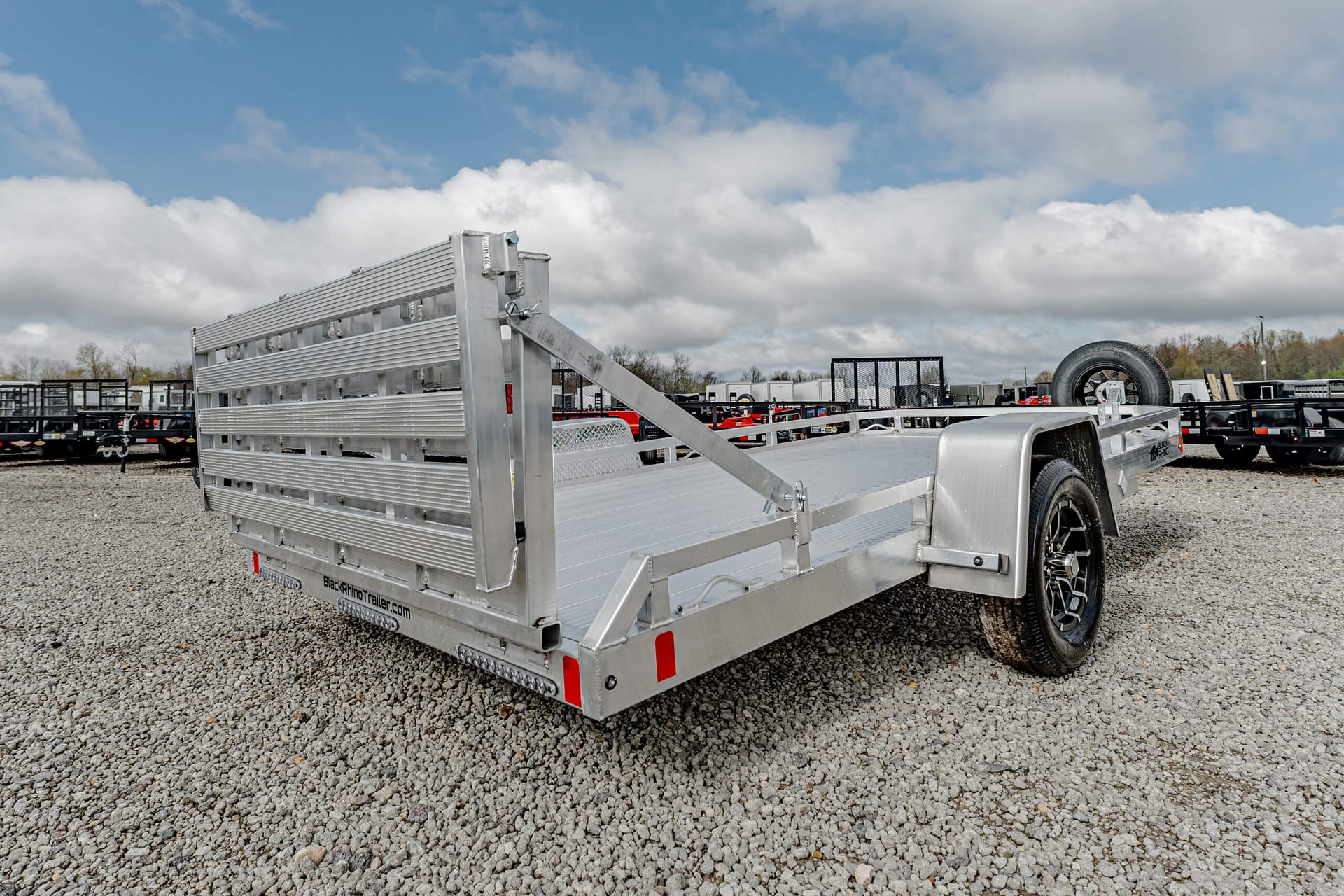 Silver aluminum flatbed trailer with ramp gate, parked on a gravel lot under a cloudy sky