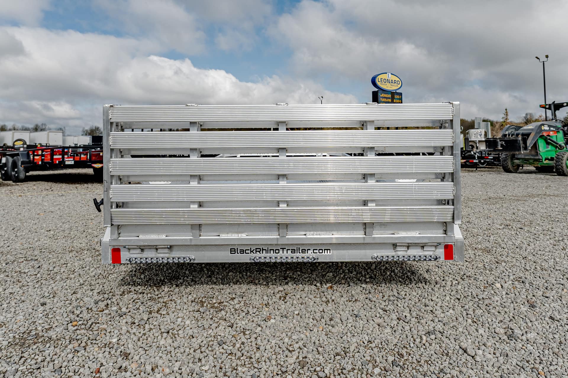 Rear view of a silver aluminum trailer with slatted ramp gate, on a gravel lot at a trailer dealership; URL BlackRhinoTrailer.com visible on bumper.