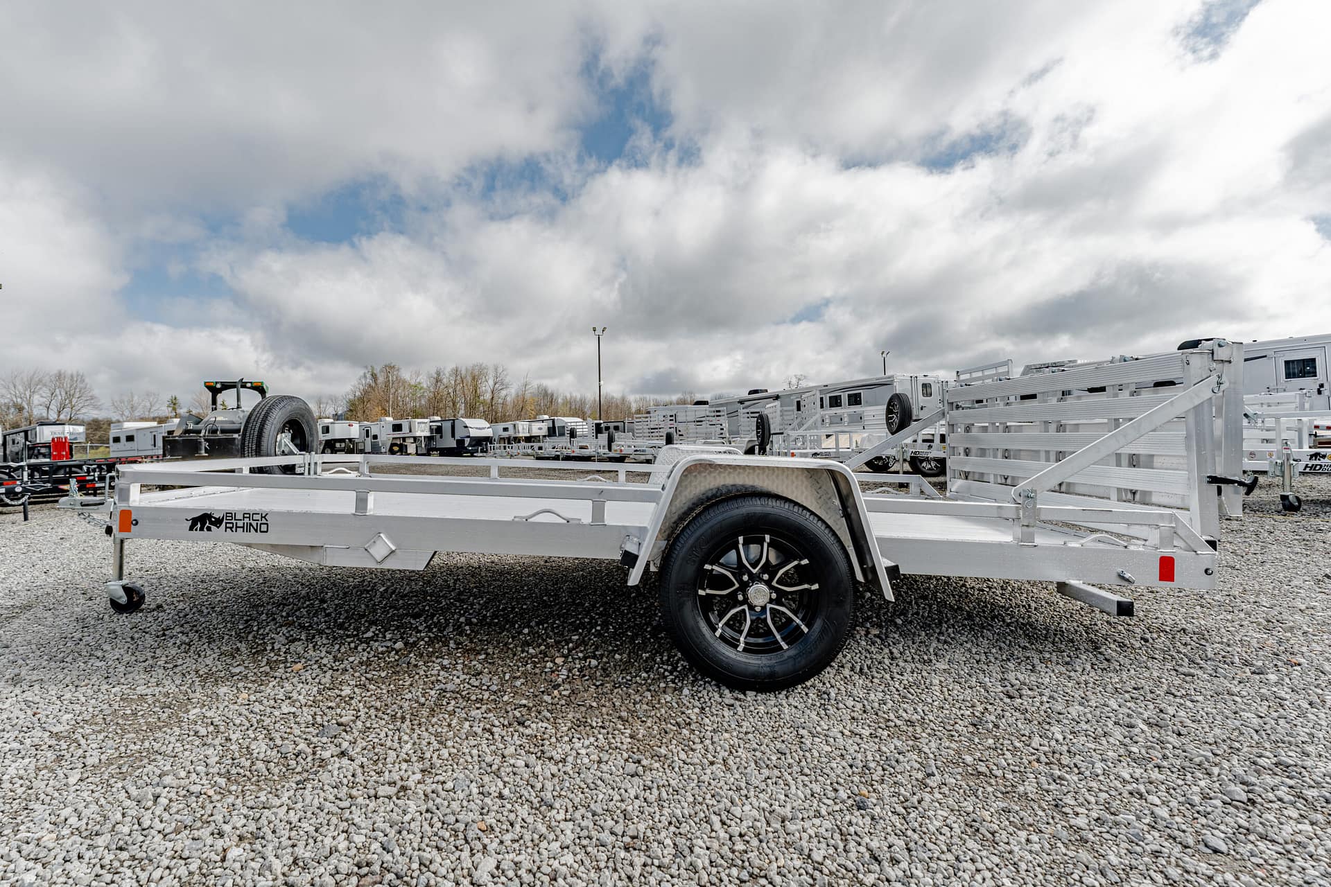 Aluminum utility trailer with a single axle and black-rimmed wheel on a gravel lot, logo reads 'Black Rhino' at the front corner, many trailers in background.