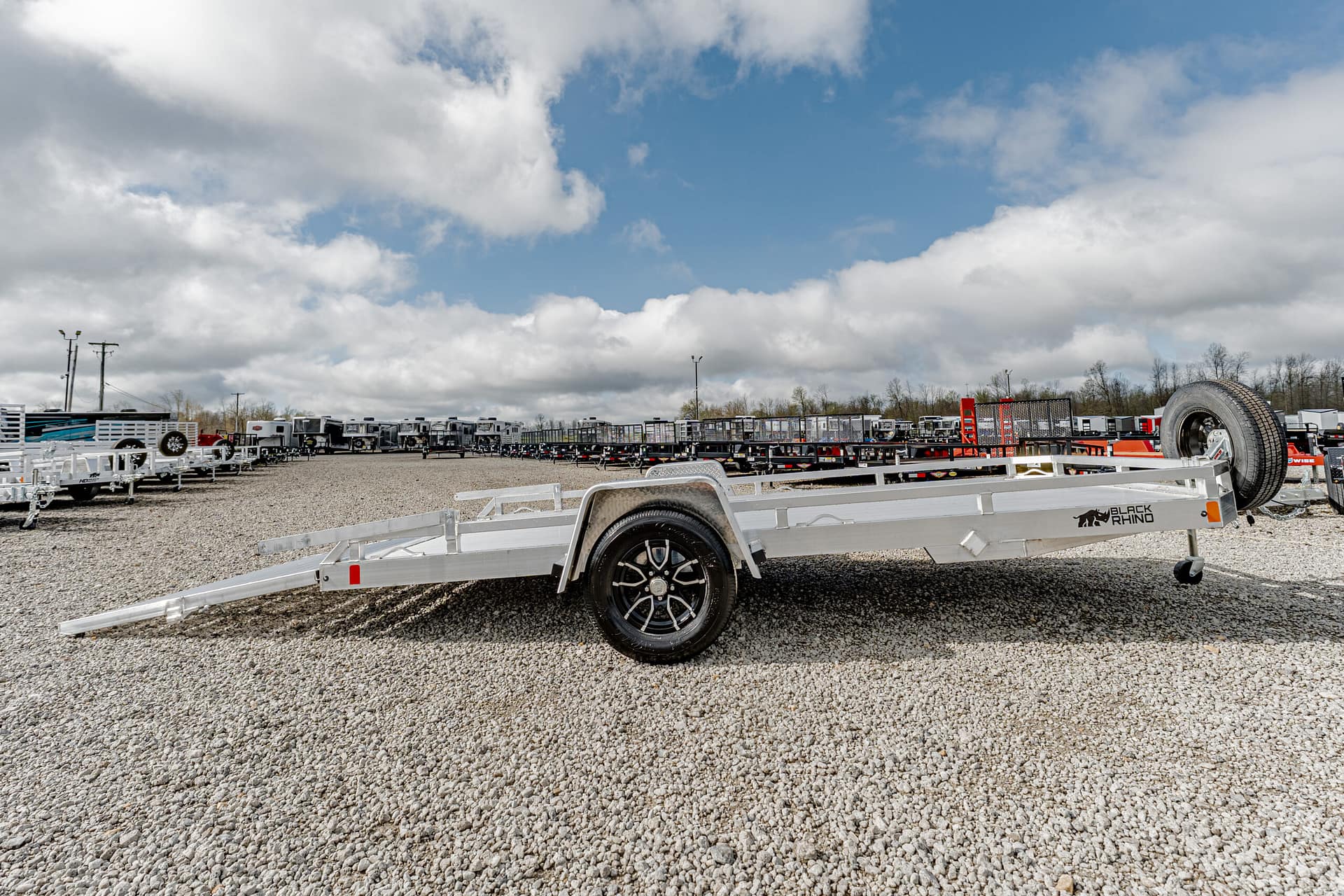 Long aluminum car trailer on a gravel lot with a front ramp lowered, single axle wheel, and a spare tire mounted at the rear (Black Rhino logo).