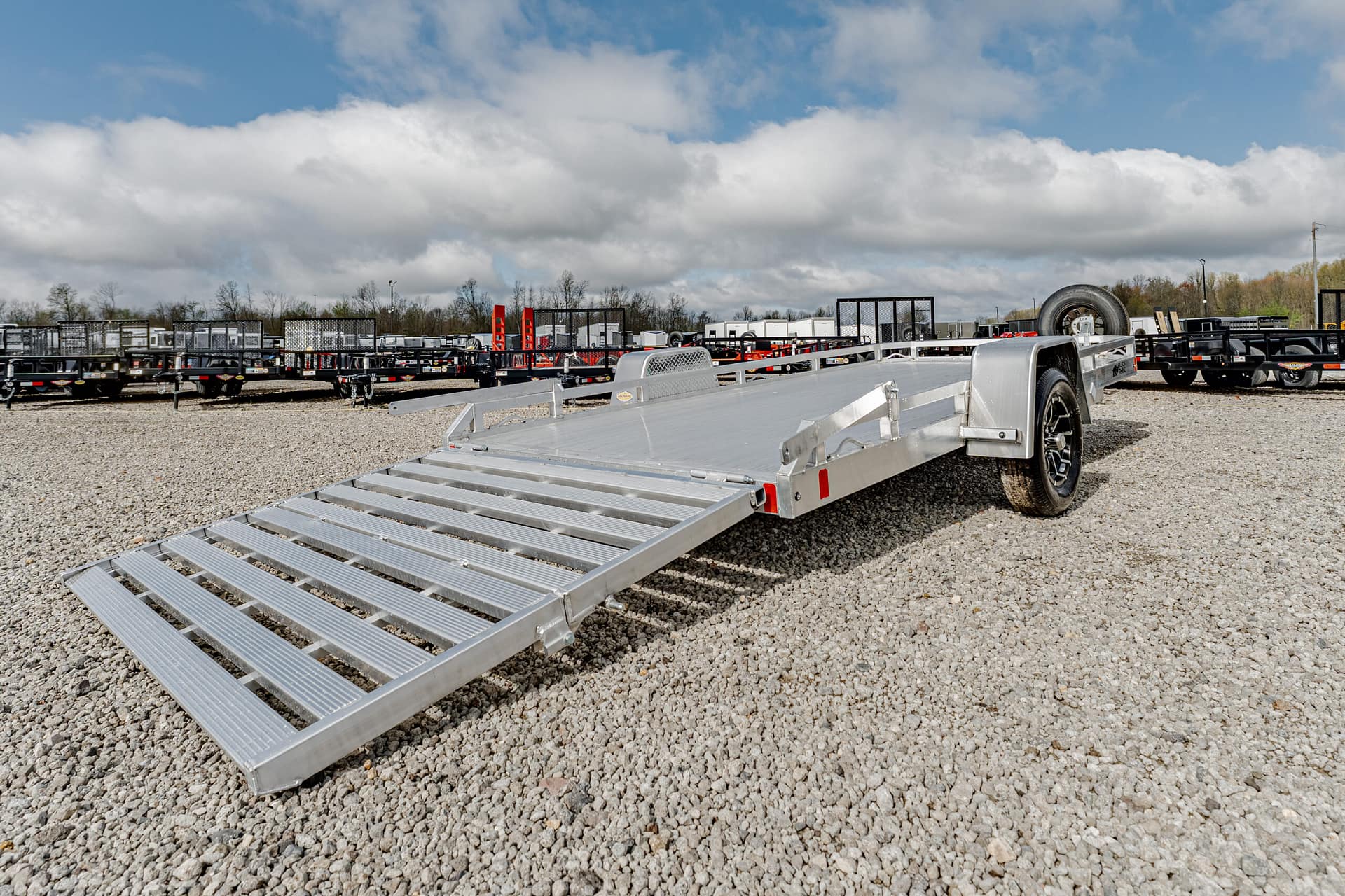 Aluminum equipment trailer with a long loading ramp extended on a gravel lot, with other trailers in the background.