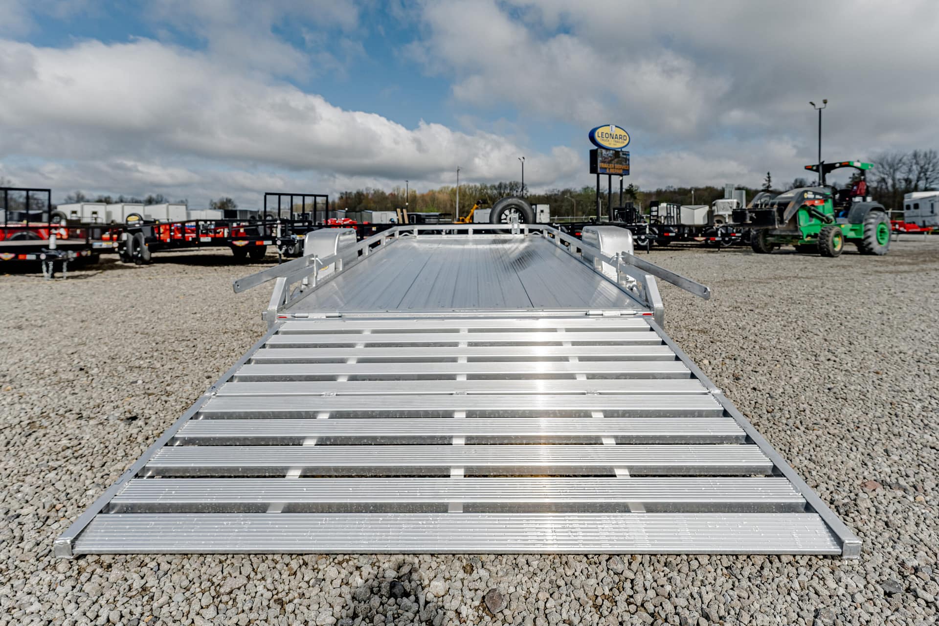 Silver aluminum flatbed trailer ramp in a gravel lot with other trailers and equipment in the background