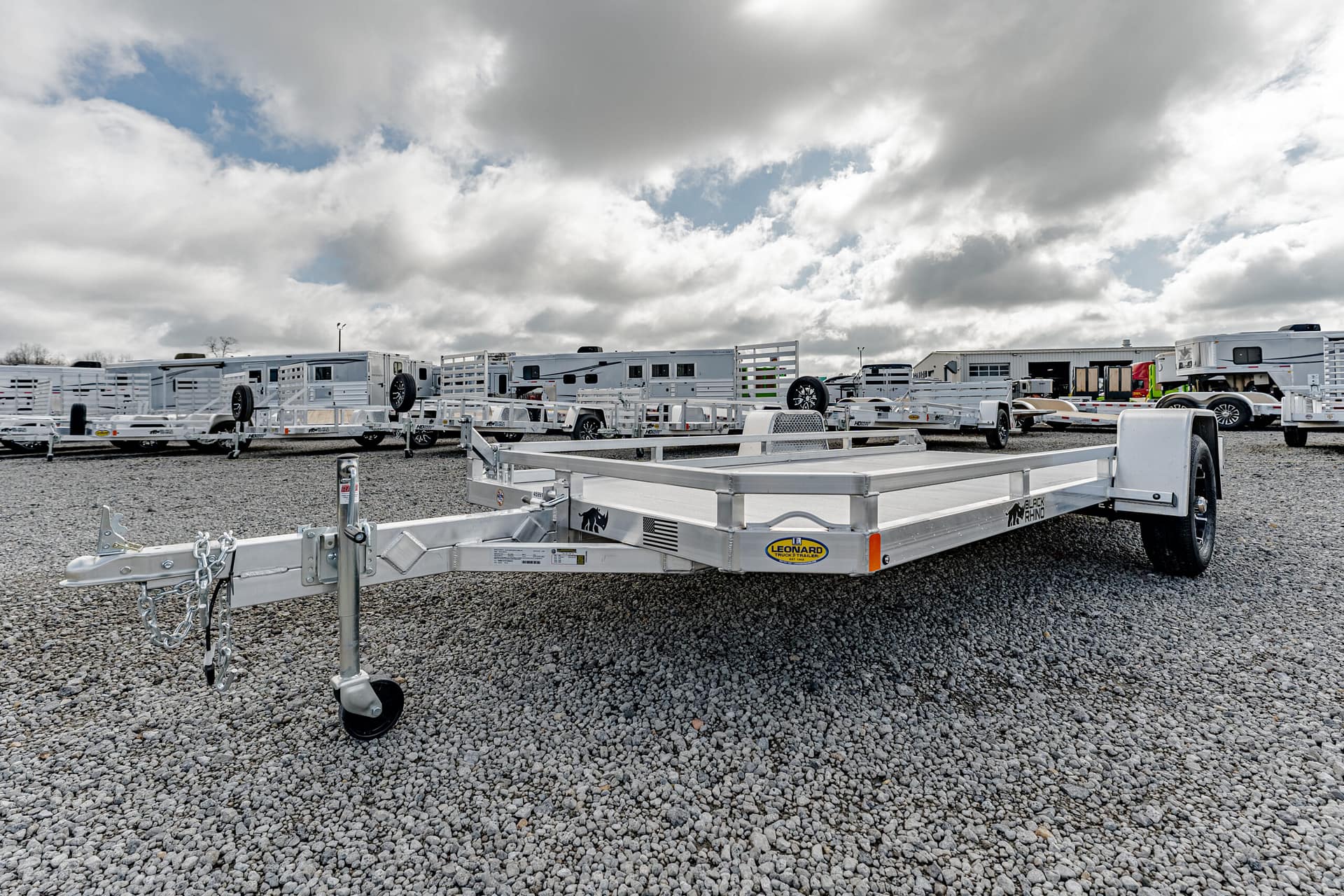 Row of white utility and horse trailers parked on a gravel lot under a cloudy sky, a trailer hitch in the foreground.