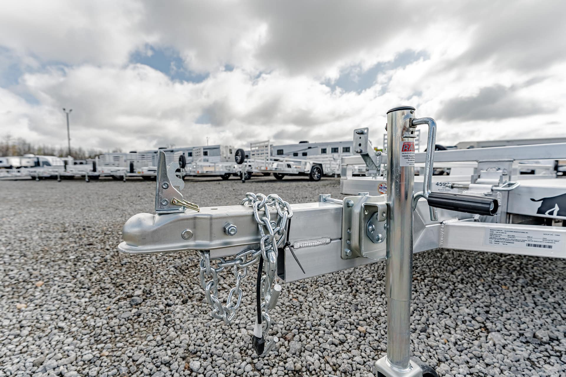 Close-up of a metal trailer hitch with chains and lock, on a gravel lot with white trailers in background under cloudy sky.