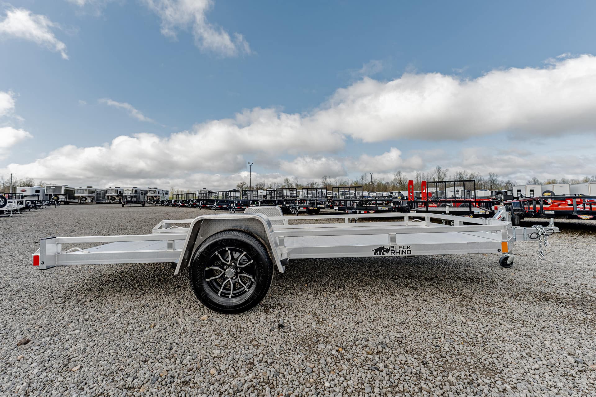 Silver utility trailer with large wheel and spare tire on a gravel lot, row of trailers in the background, under a blue sky with clouds to the horizon.
