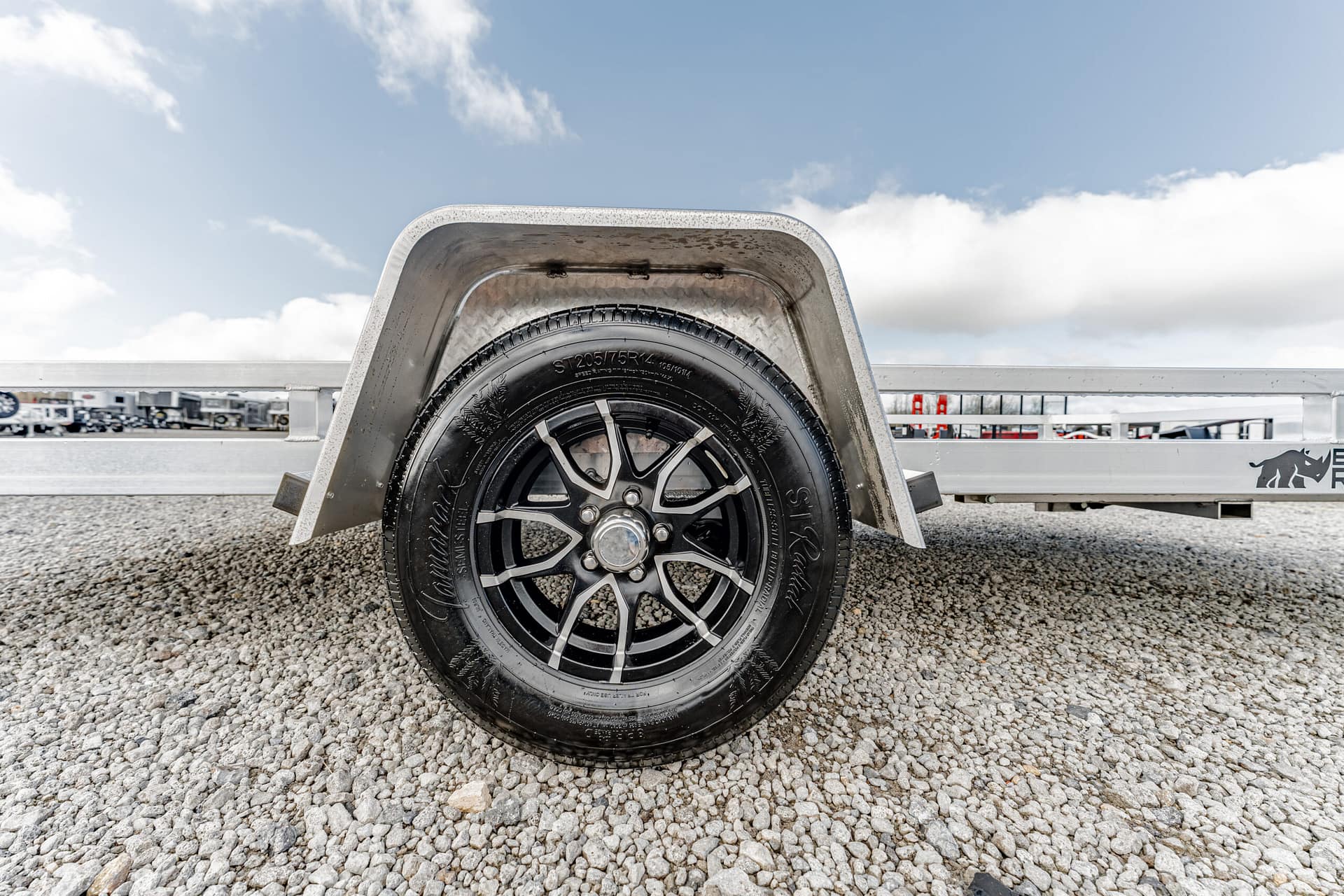 Close-up of a trailer wheel inside a metal fender on a gravel surface with a blue sky above.