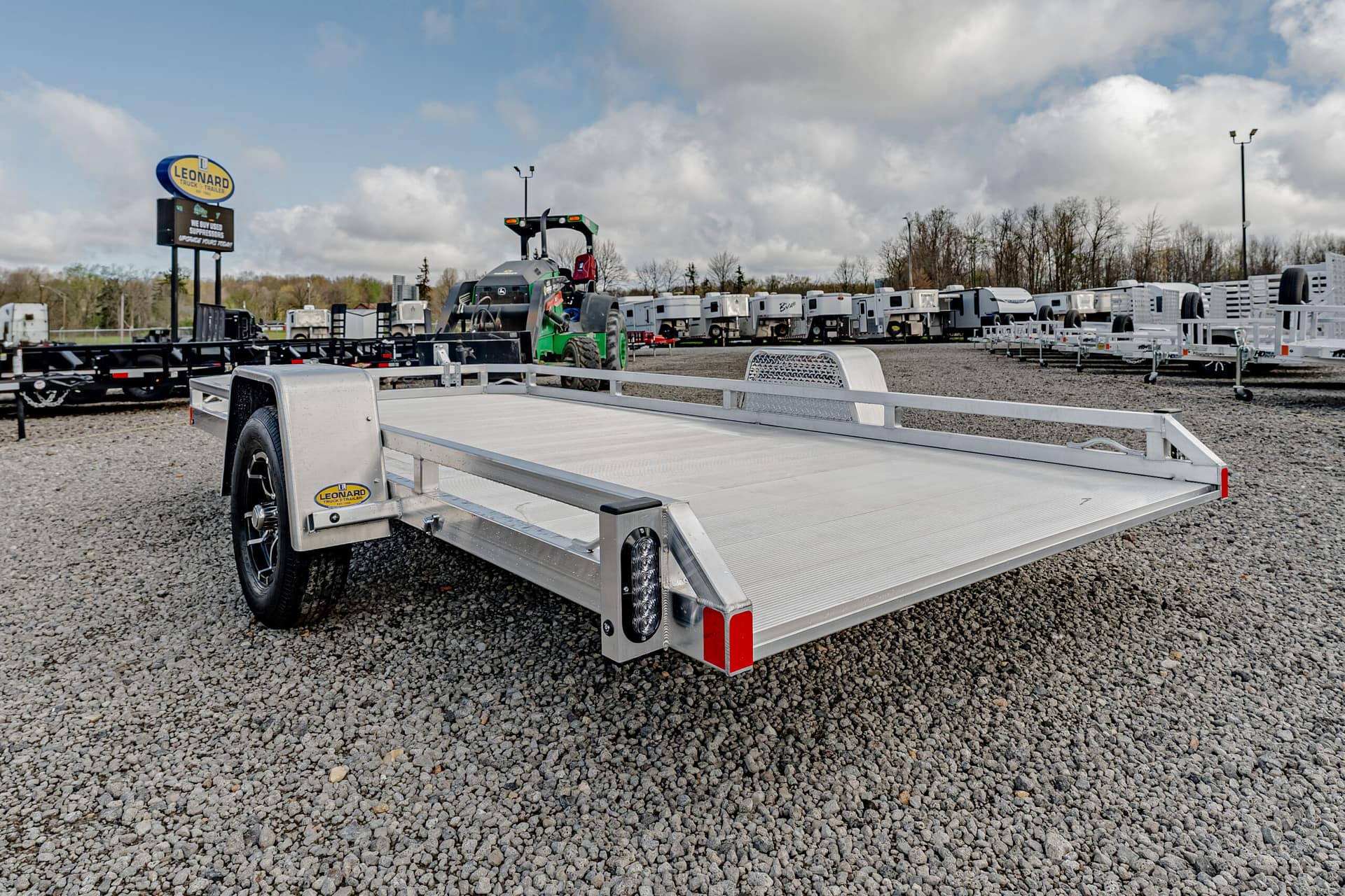 Silver flatbed trailer in the foreground at a Leonard Truck & Trailer lot, with rows of trailers and equipment under a cloudy sky in the background.