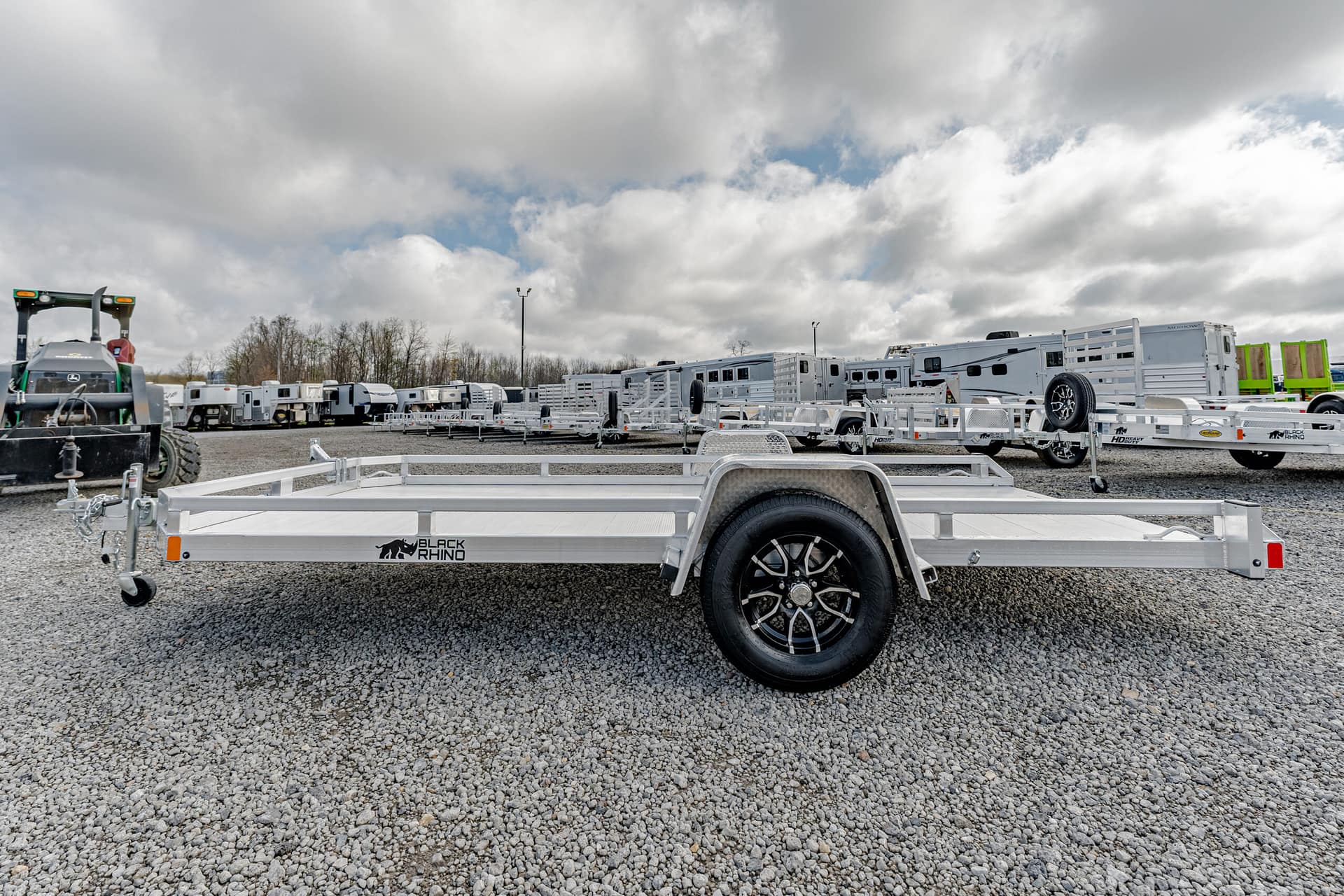 Row of aluminum trailers parked in a gravel lot under a cloudy sky; foreground shows a close-up of a Black Rhino trailer wheel.
