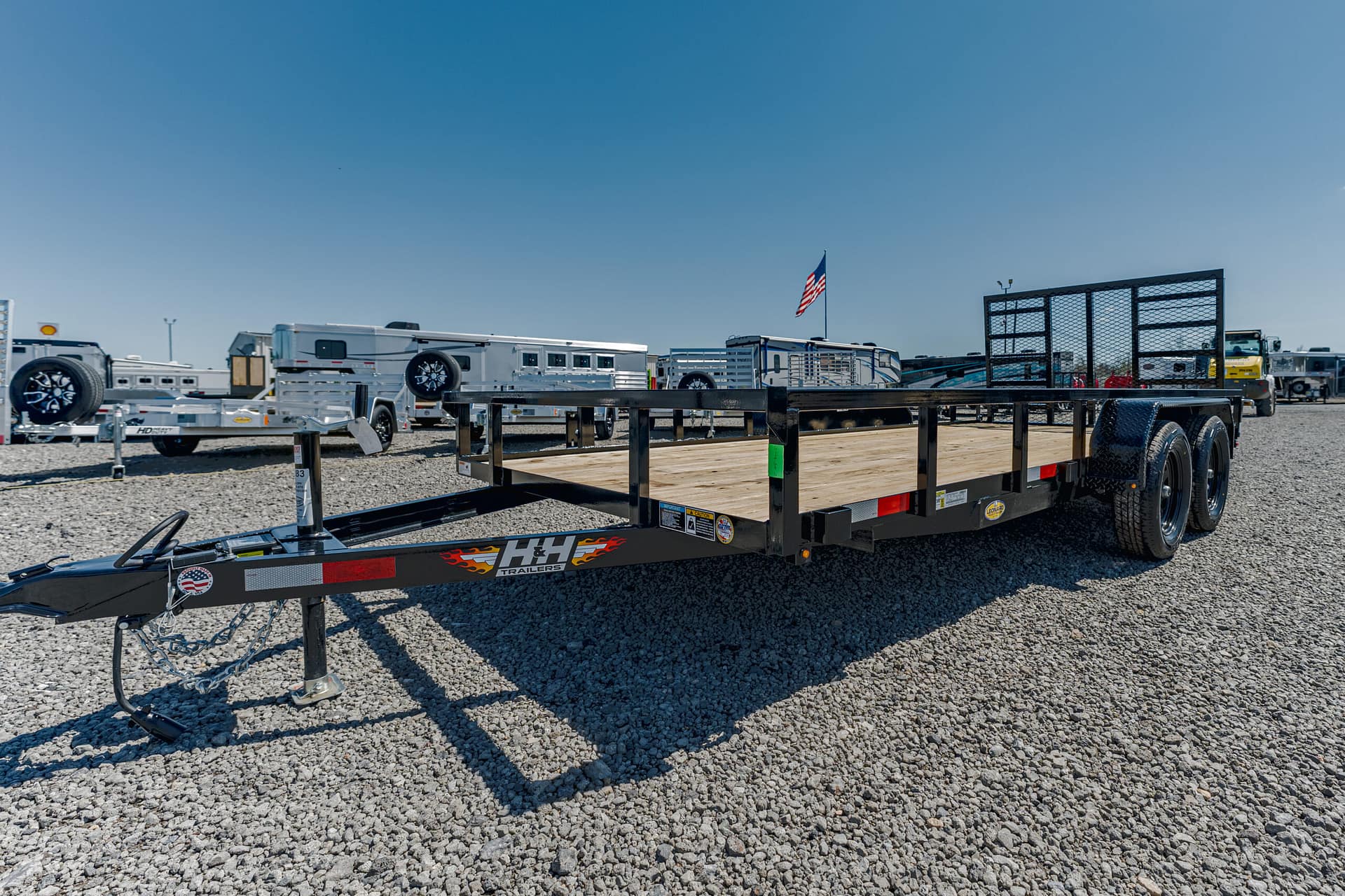Black flatbed trailer with a wooden deck in a gravel trailer yard, American flag visible in the background.