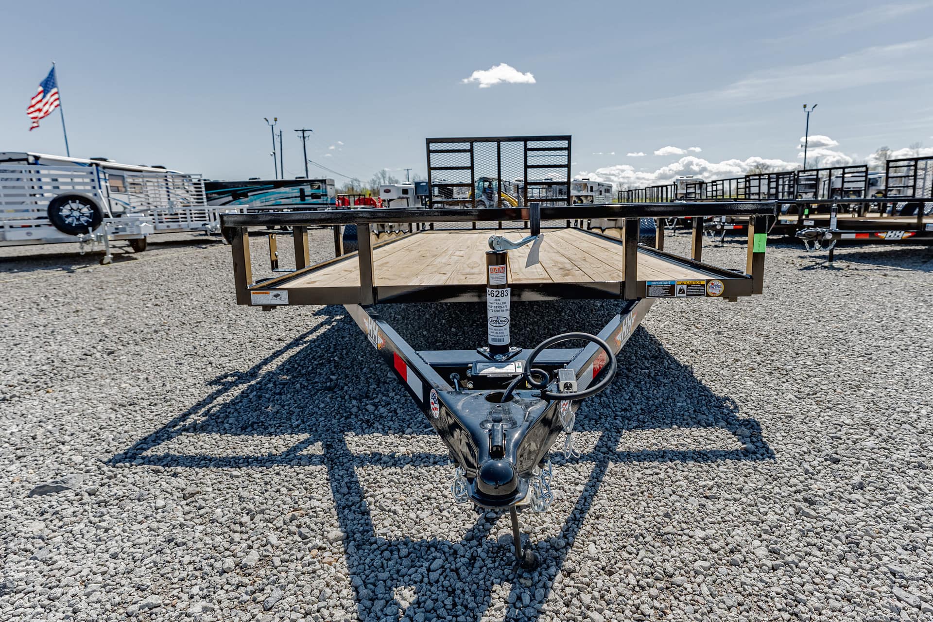 Front view of a flatbed trailer hitch on a gravel lot with other trailers in the background and an American flag on the left edge.",