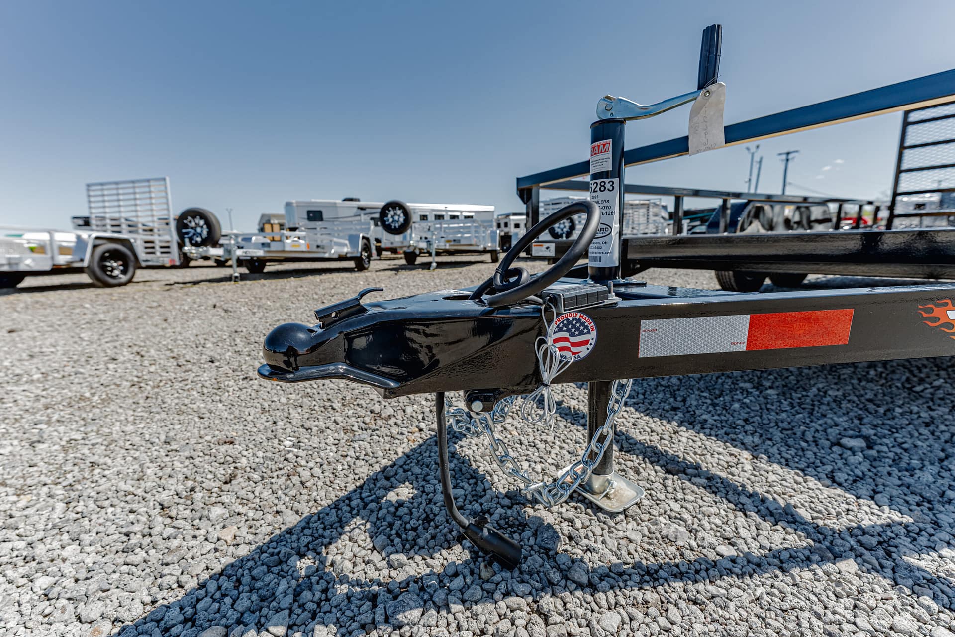 Close-up of a trailer hitch on a gravel lot with other trailers in the background under a clear blue sky, conveying an outdoor trailer yard.