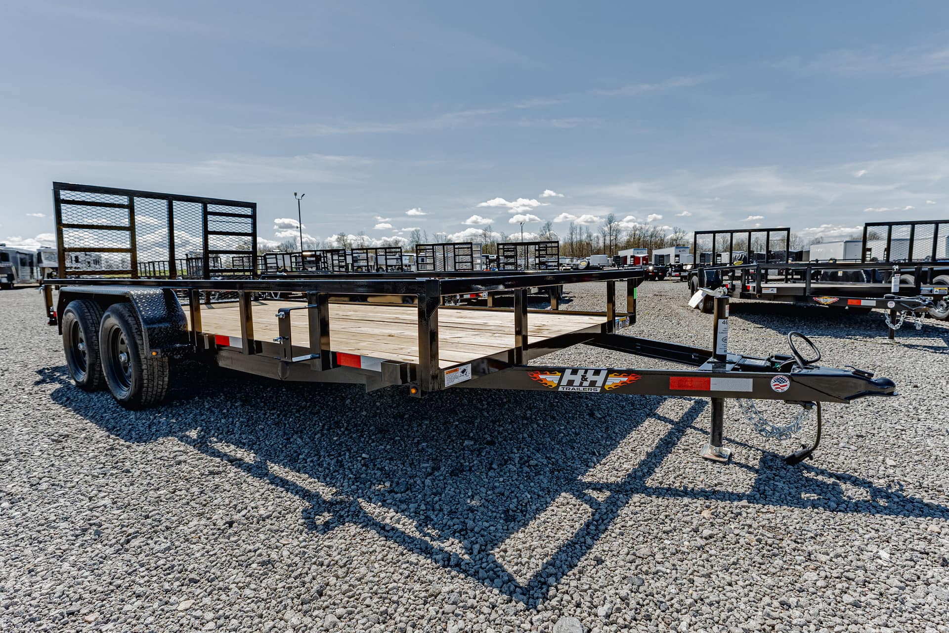 Flatbed trailer with a wooden deck and side rails parked on a gravel lot, with more trailers in the background under a blue sky.