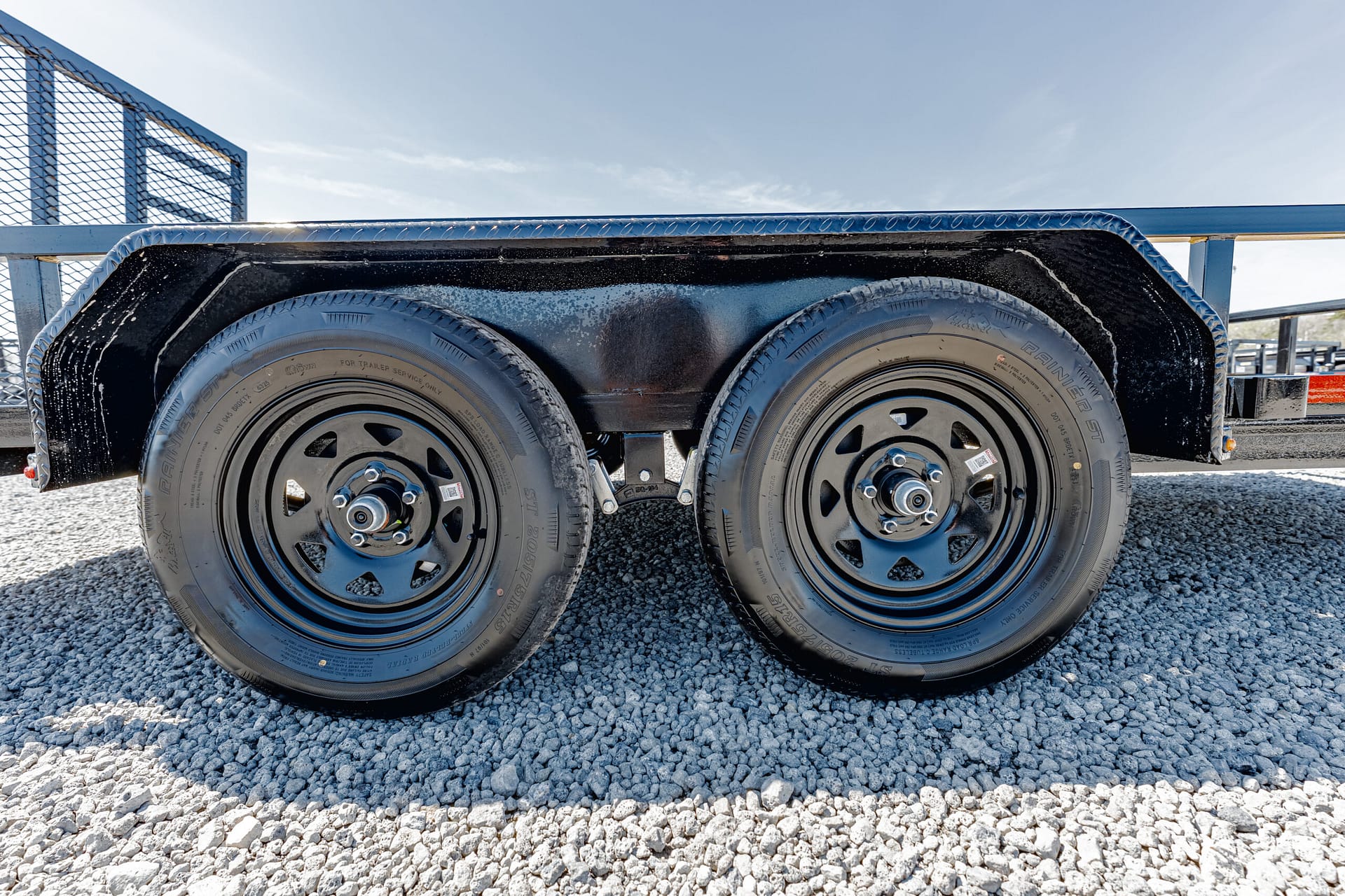 Close-up of dual trailer wheels with black fenders on a gravel surface and blue sky above, showing shiny lug nuts and tires.