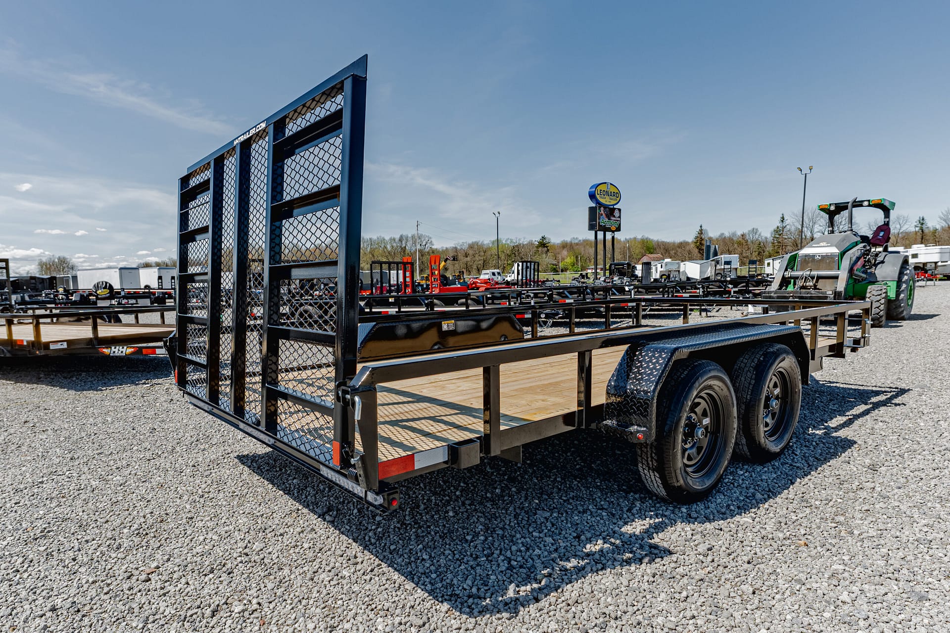 Large flatbed trailer in a gravel lot with a tall mesh ramp and dual rear wheels, clear blue sky above