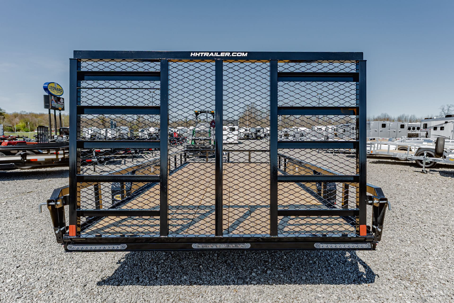 Front view of a blue metal utility trailer with diamond mesh gate, parked on gravel lot; HHtrailer.com logo on top beam; other trailers visible in background.