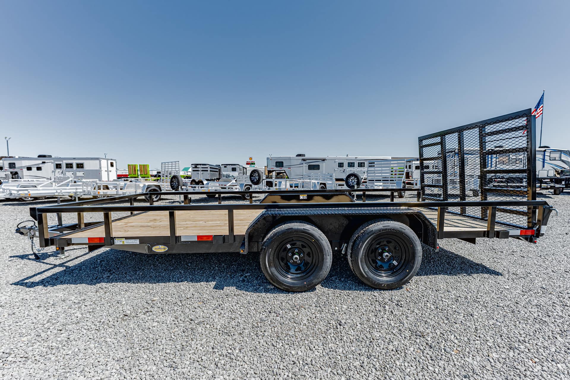 Flatbed trailer on a gravel lot with other trailers in the background, a fenced gate on the right, and an American flag in the distance under a clear blue sky.