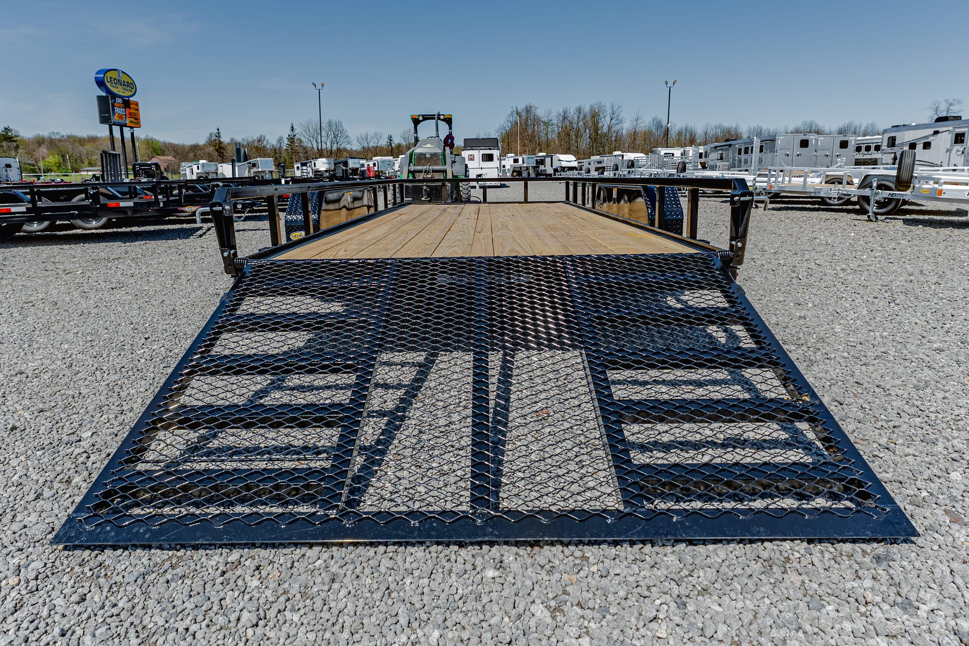 Front view of a black metal utility trailer with a wooden deck in a gravel lot, with many trailers and RVs in the background.