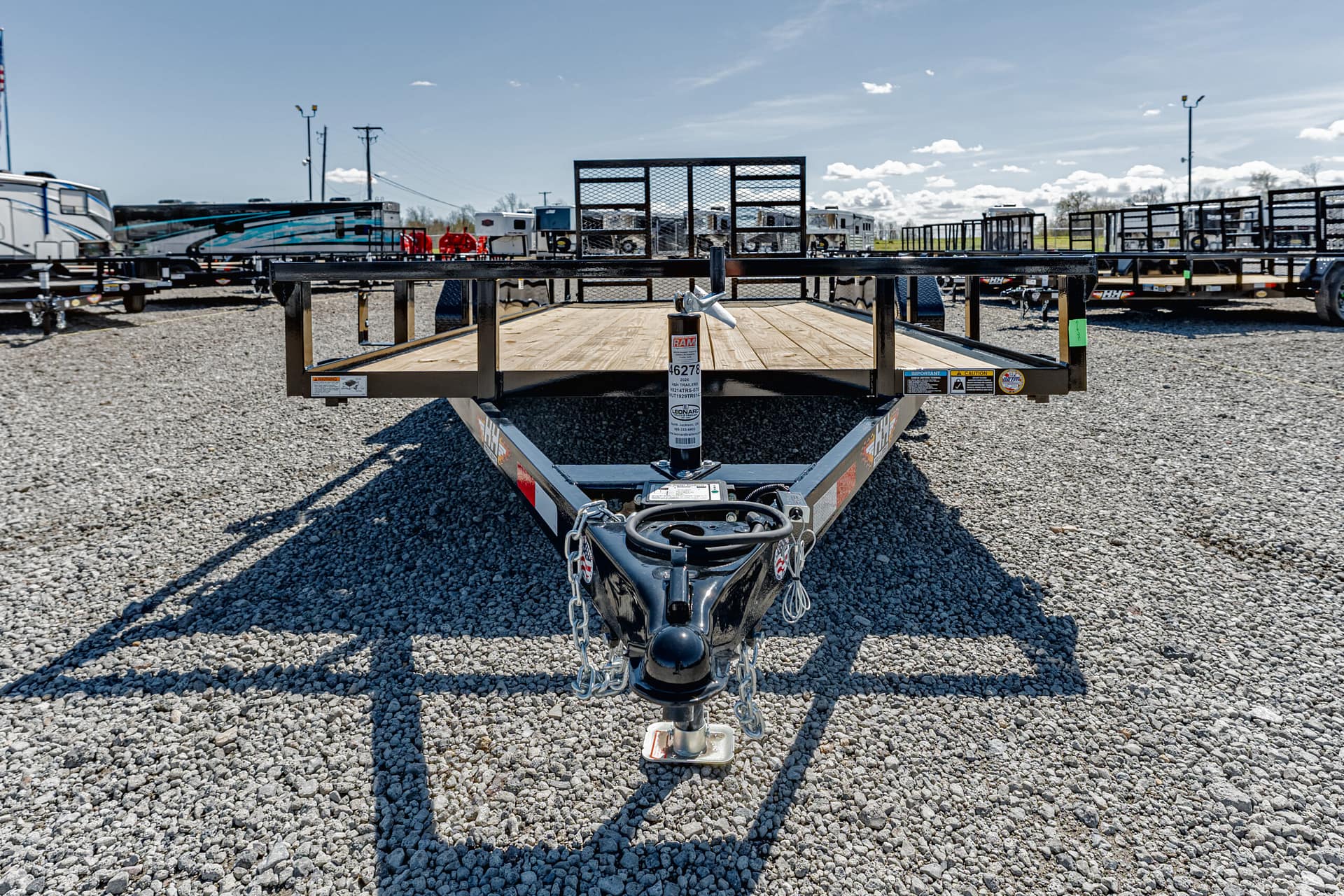 Front view of a flatbed trailer hitch on a gravel lot with other trailers in the background under a blue sky.