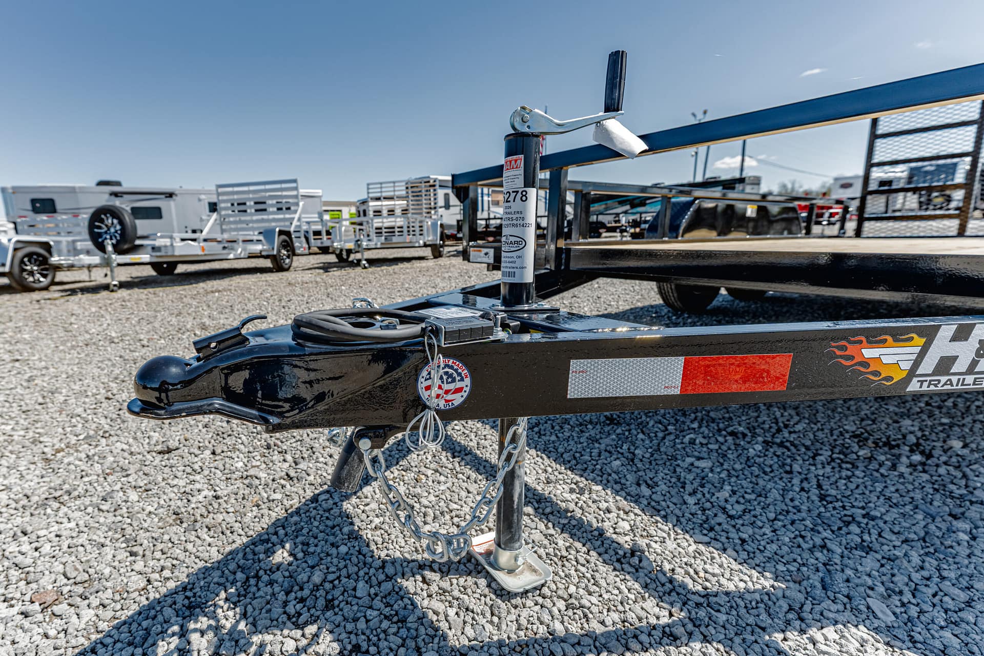 Close-up of a metal trailer hitch with chains on a black boat trailer in a gravel lot, other trailers in the background under a blue sky.