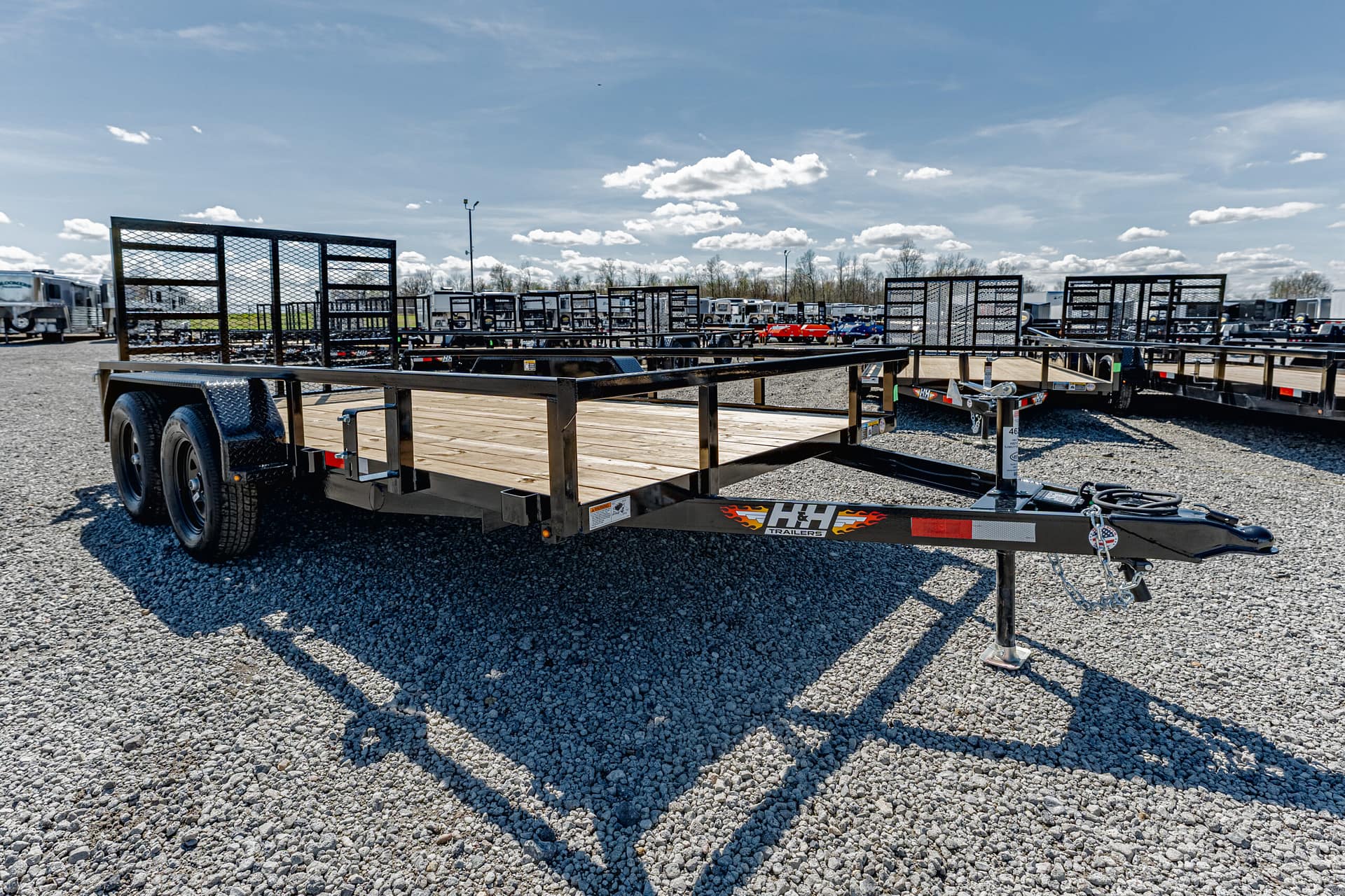 Front view of a black flatbed trailer with a wooden deck in a gravel lot, with other trailers in the background under a blue sky.