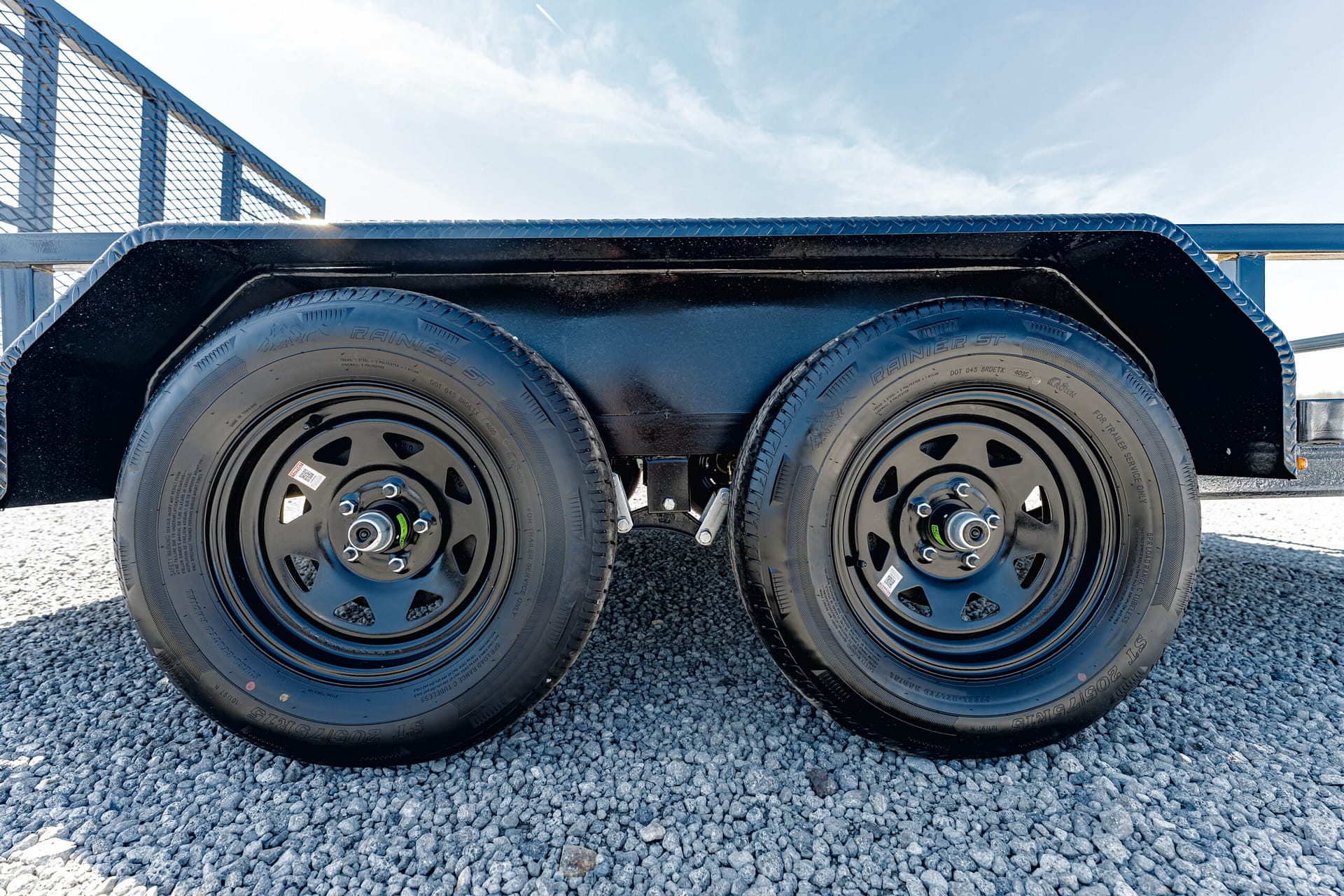 Dual-axle trailer with black fenders and two large tires on a gravel surface under a clear blue sky