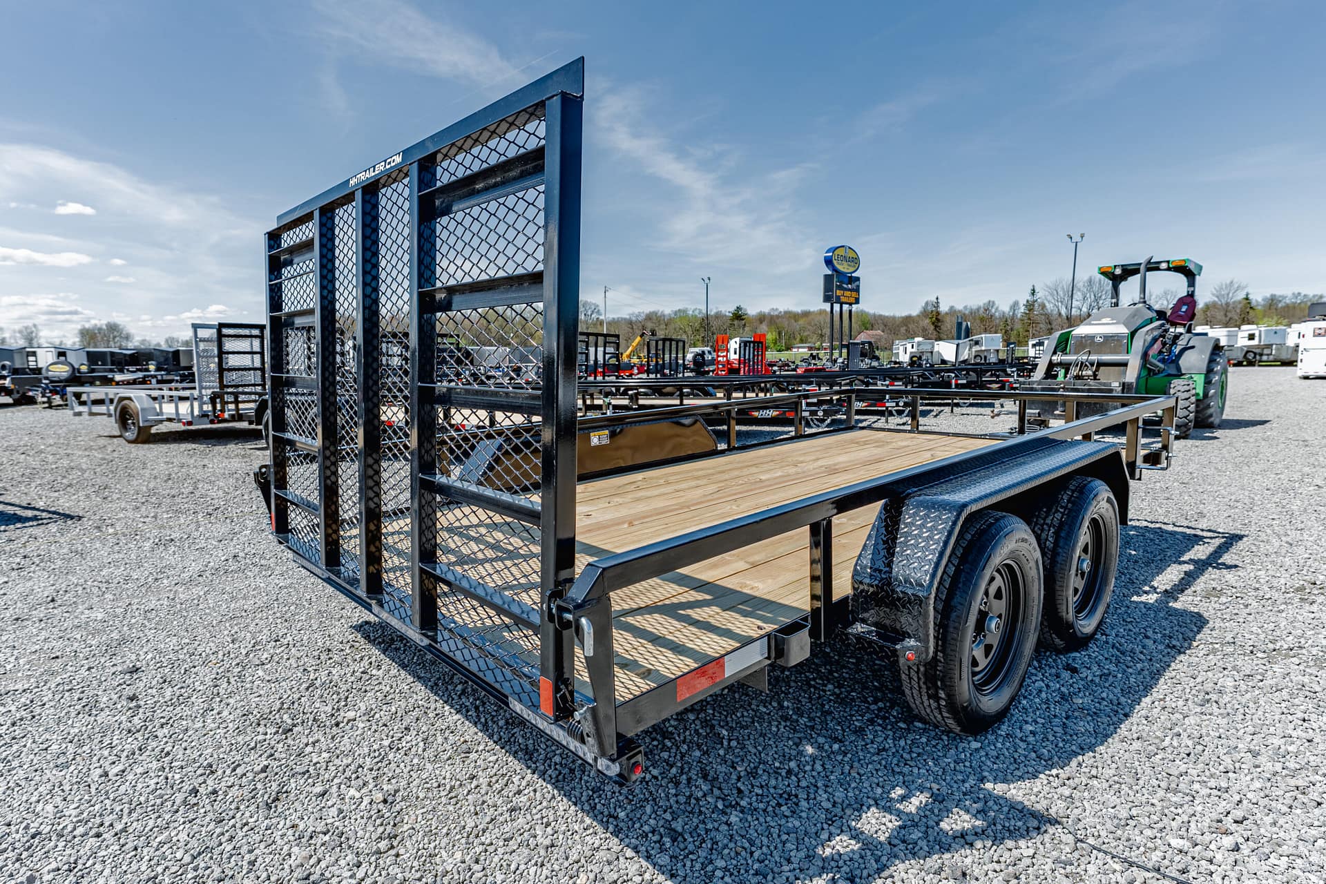 Flatbed trailer with a metal mesh guard in the foreground on a gravel lot, other trailers and equipment visible in the background under a blue sky.