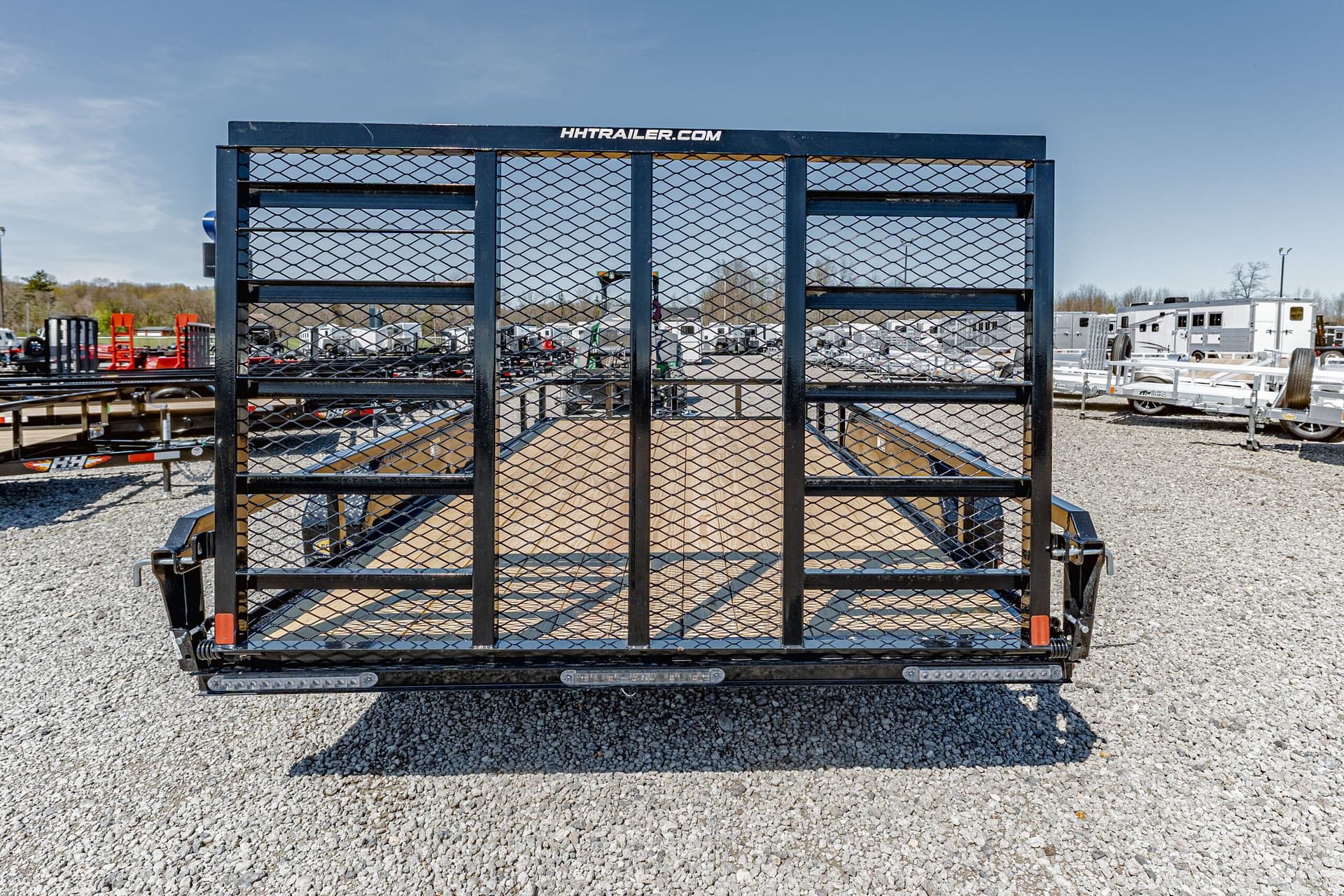 Front view of a black utility trailer with diamond-mesh sides on a gravel lot, with other trailers and RVs in the background.
