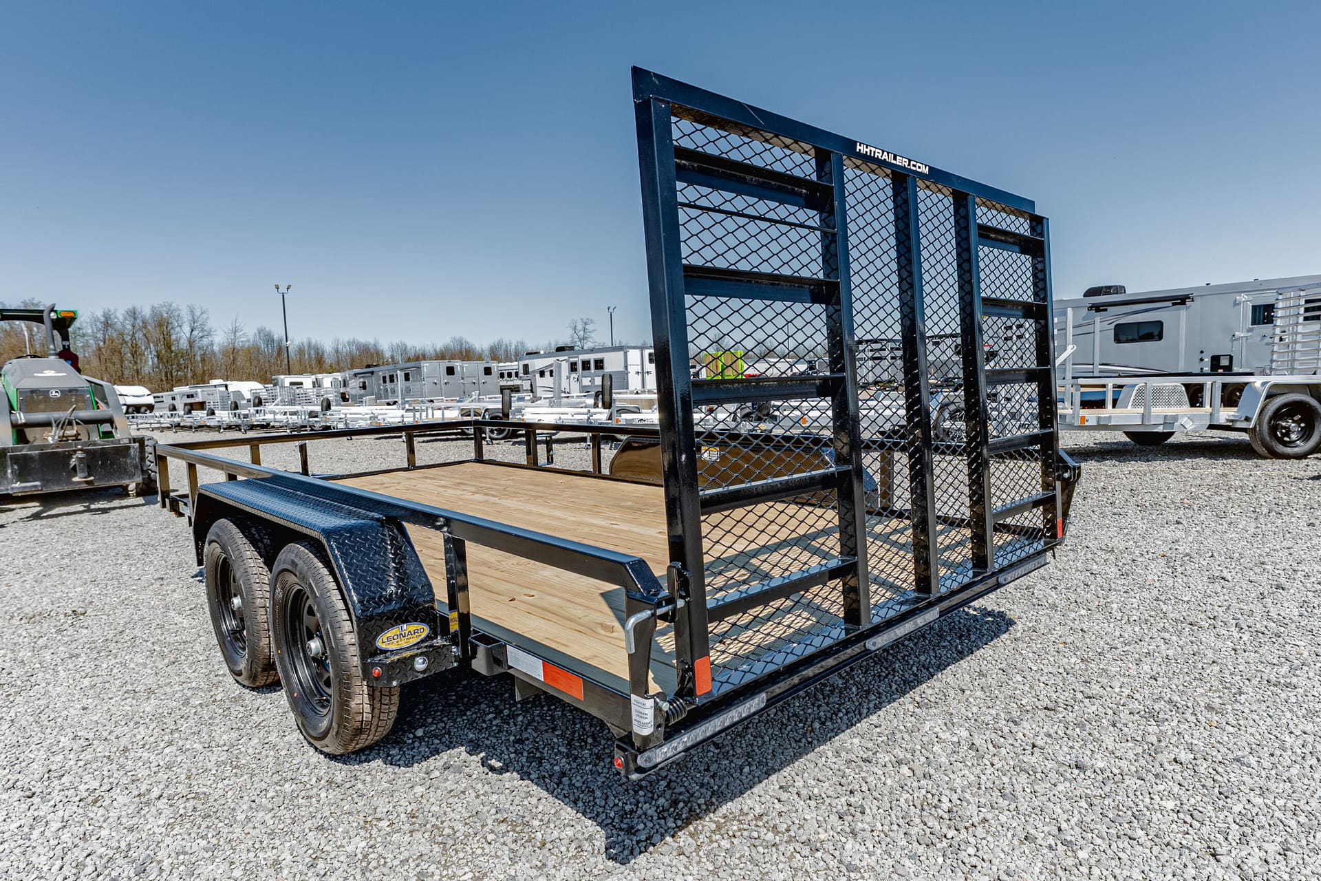 Black utility trailer with a wooden deck and mesh gate in a gravel lot, with other trailers and RVs in the background on a sunny day.