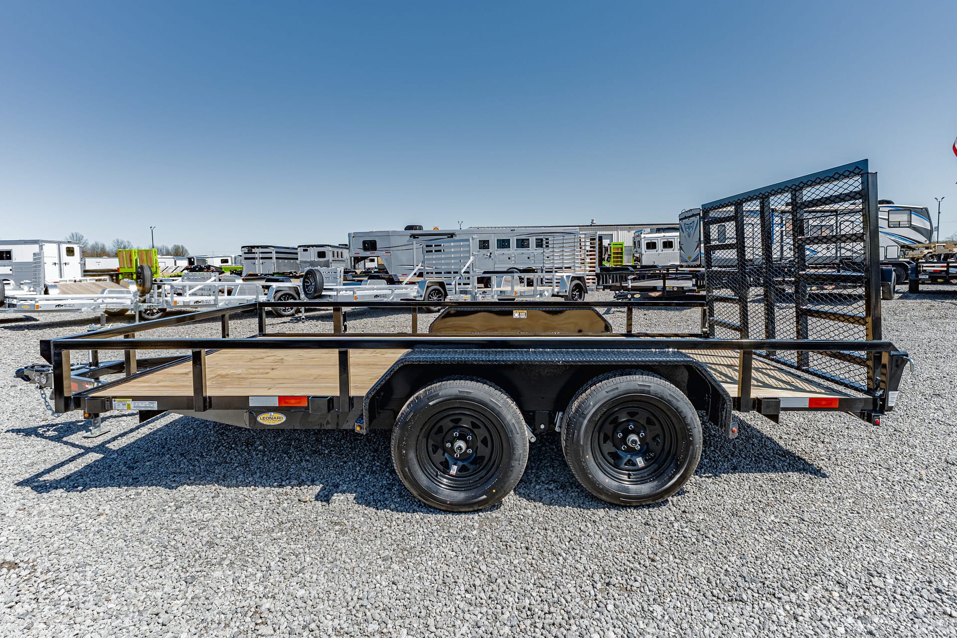 Wide view of a trailer yard filled with multiple flatbed and horse trailers on a gravel lot under a clear blue sky.
