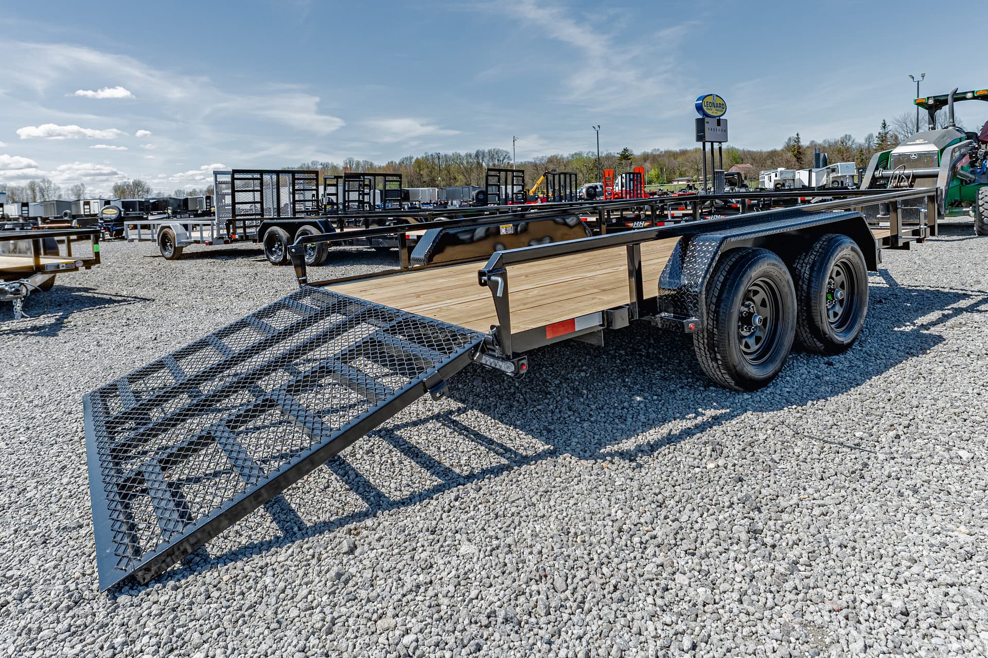 Wide view of a trailer yard: multiple flatbed trailers with ramps on a gravel lot under a blue sky.