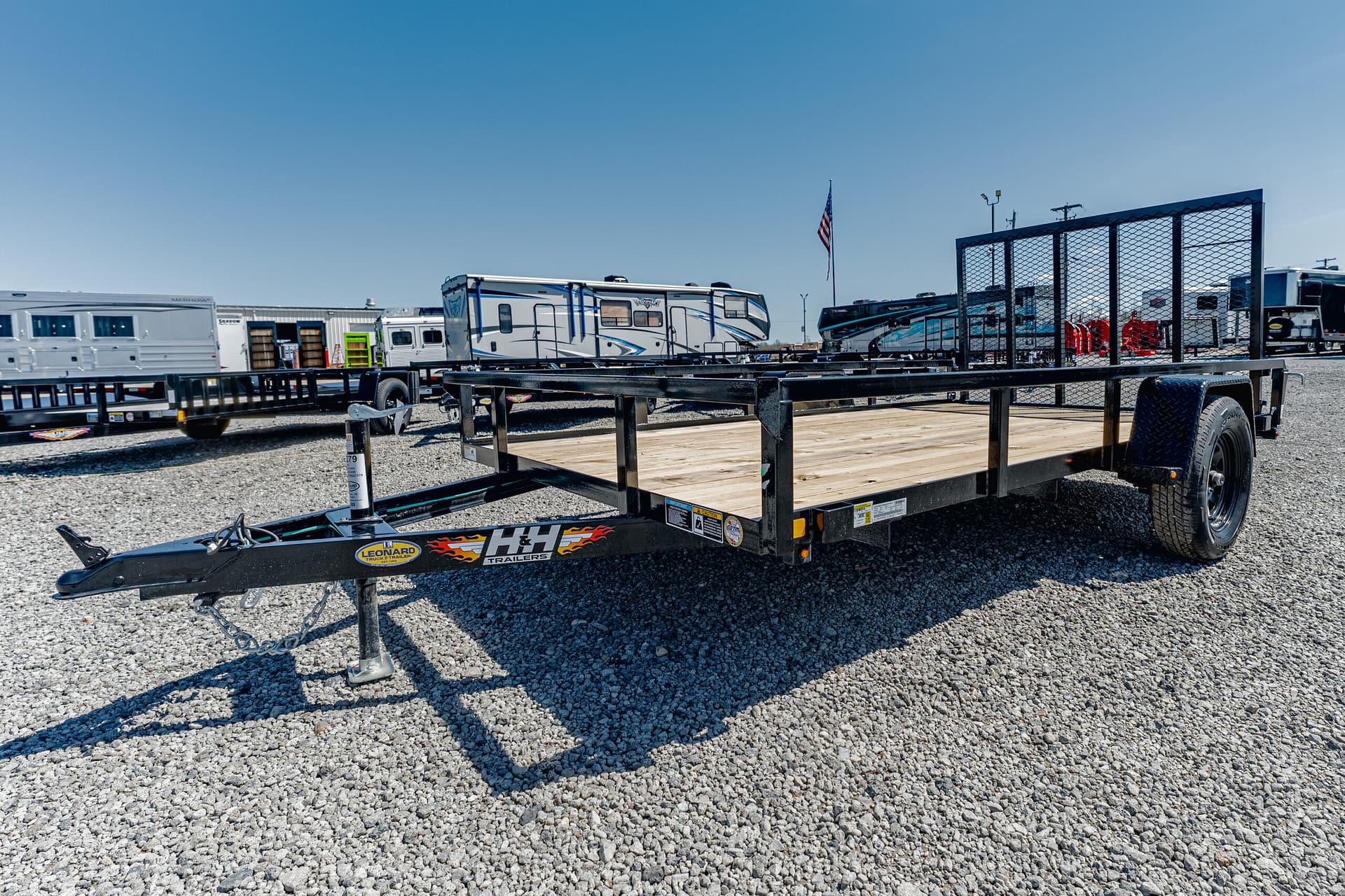 Black metal-framed flatbed trailer with a wooden deck, side rails, and a single axle, parked on a gravel lot with other trailers nearby; hitch and jack stand at the front, branding stickers visible.