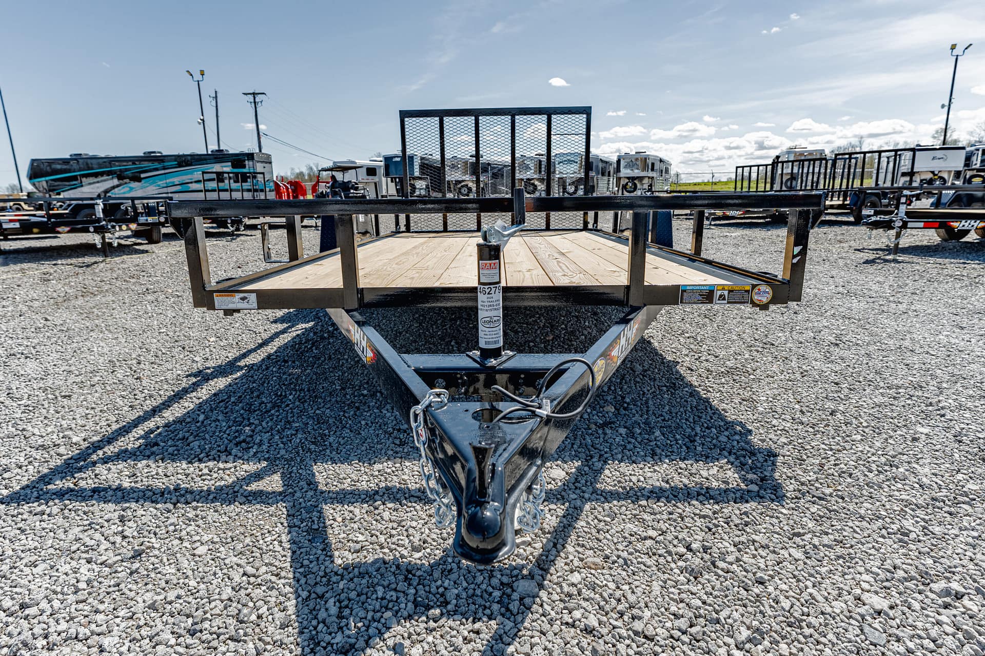 Front view of a black metal flatbed trailer with a wooden deck on a gravel lot, with RVs and trailers in the background.