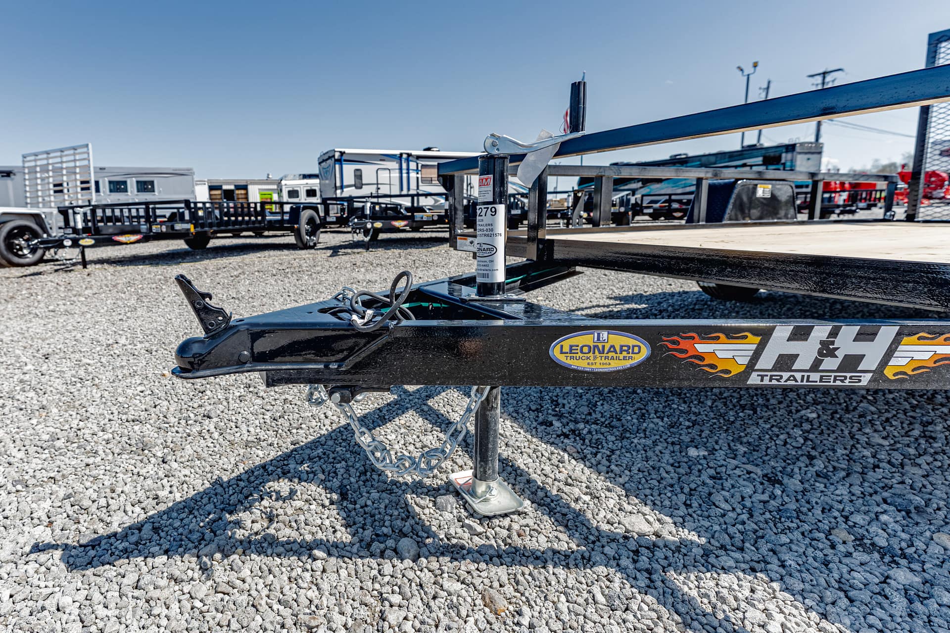Close-up of a black trailer tongue with Leonard and H&H Trailers branding, safety chain, and hitch on a gravel lot.
