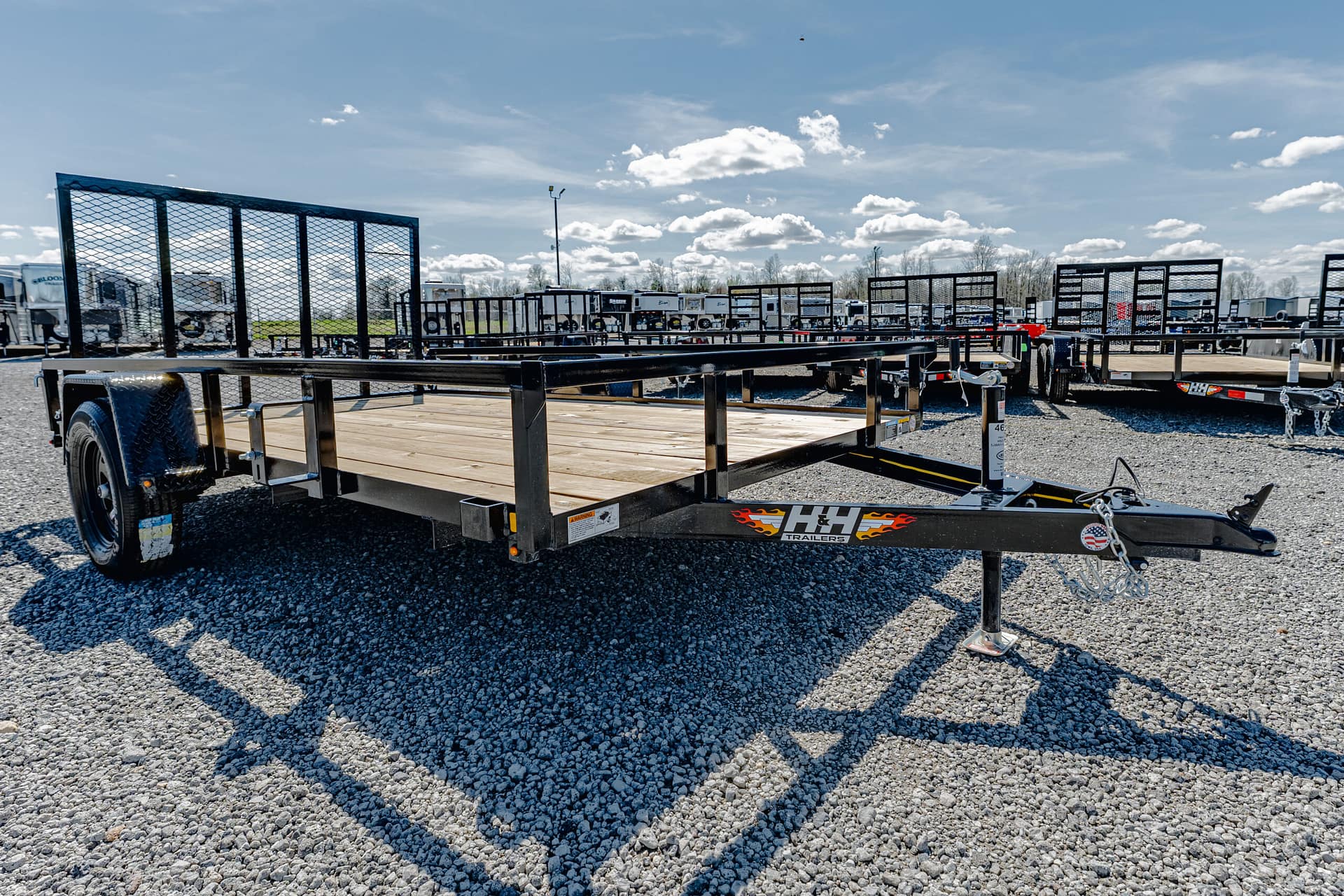 Black flatbed trailer with a wooden deck and side rails, parked on a gravel lot with other trailers in the background.