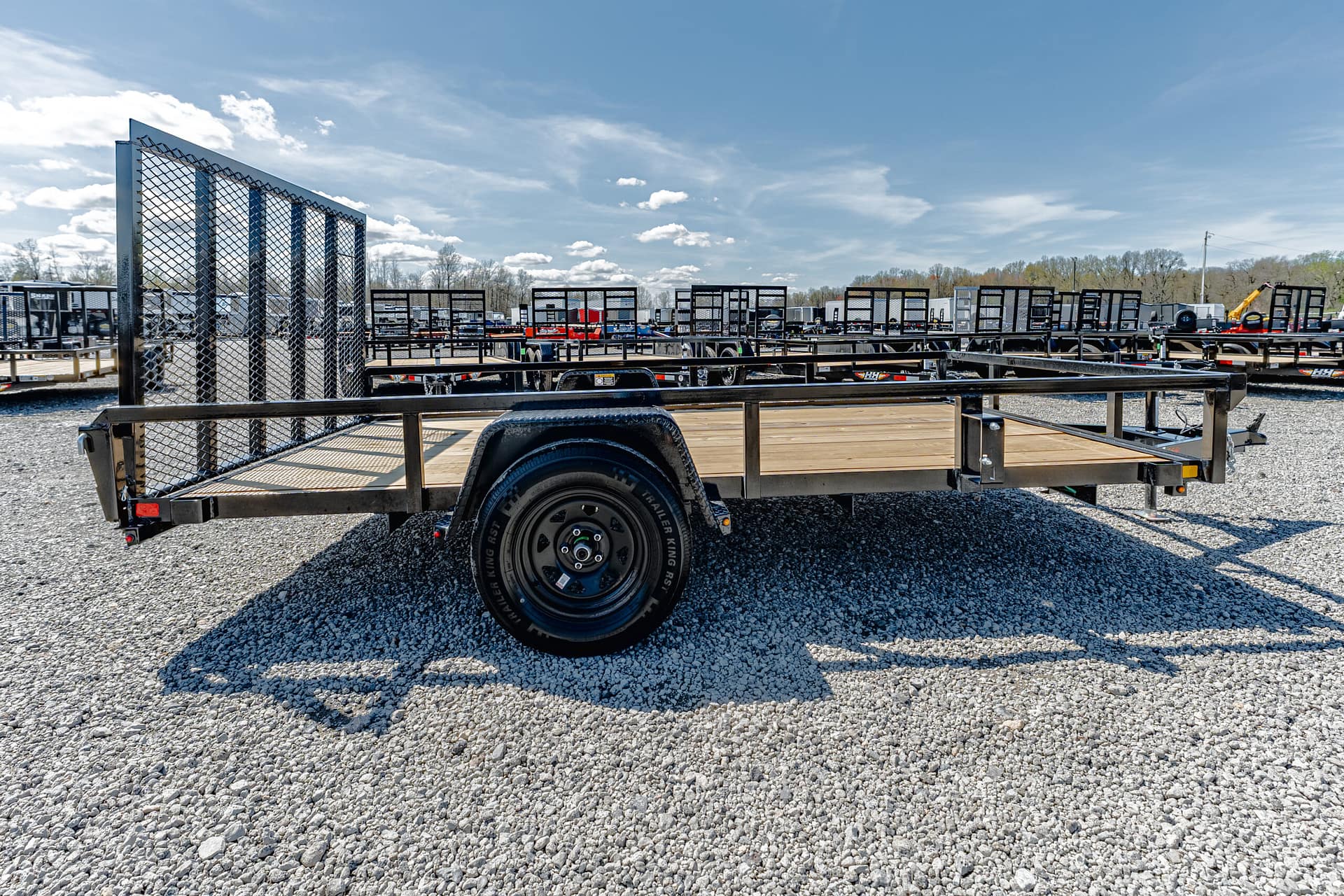 Flatbed utility trailer with wooden bed and tall metal mesh ramp, parked on gravel lot among other trailers.