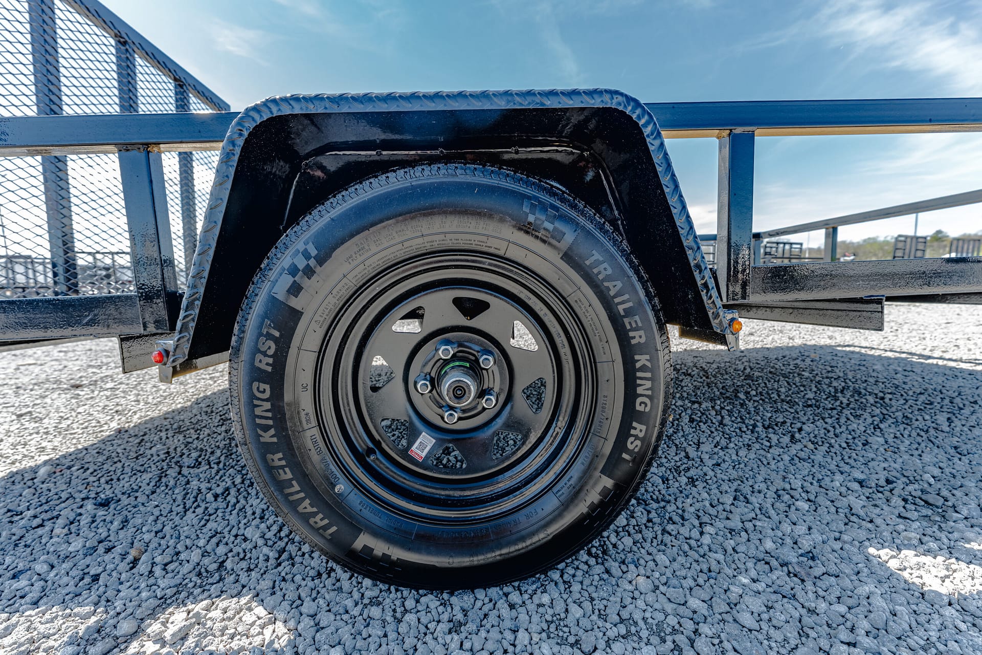Close-up of a black trailer wheel with a diamond-plate fender on a gravel surface under a blue sky.