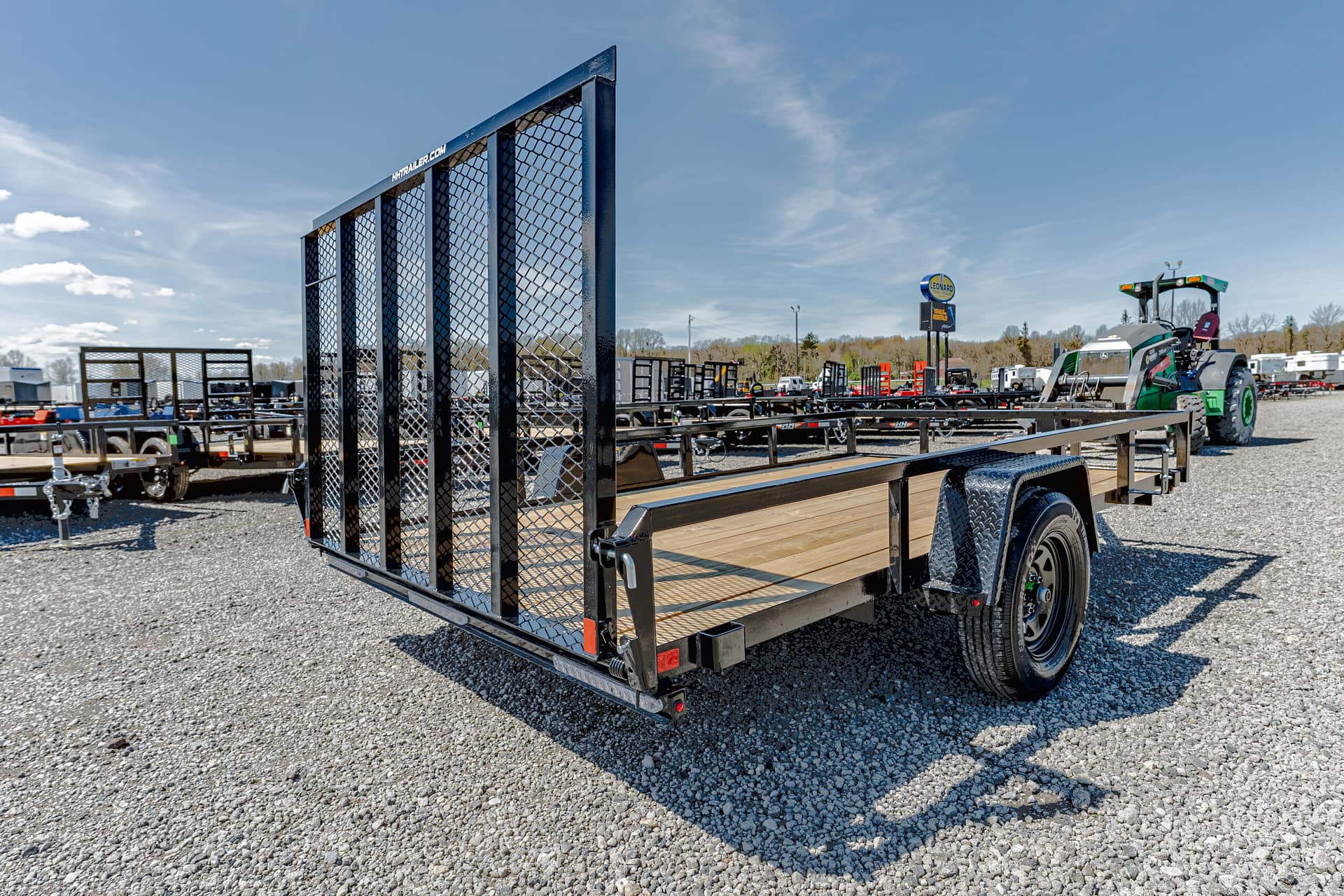Black utility trailer with a wooden deck and tall mesh ramp, parked on a gravel lot with other trailers in the background.