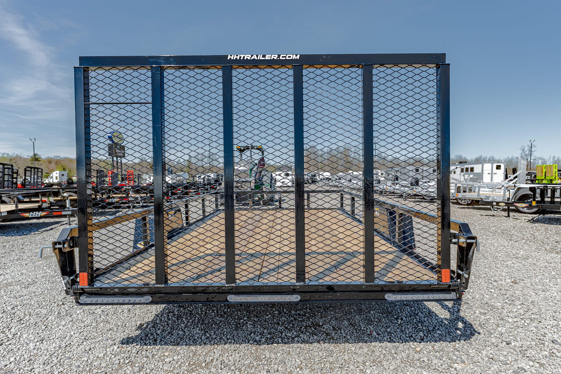 Flatbed utility trailer with a mesh gate on a gravel lot, logo HA trailer.com on the top rail, with other trailers in the background.