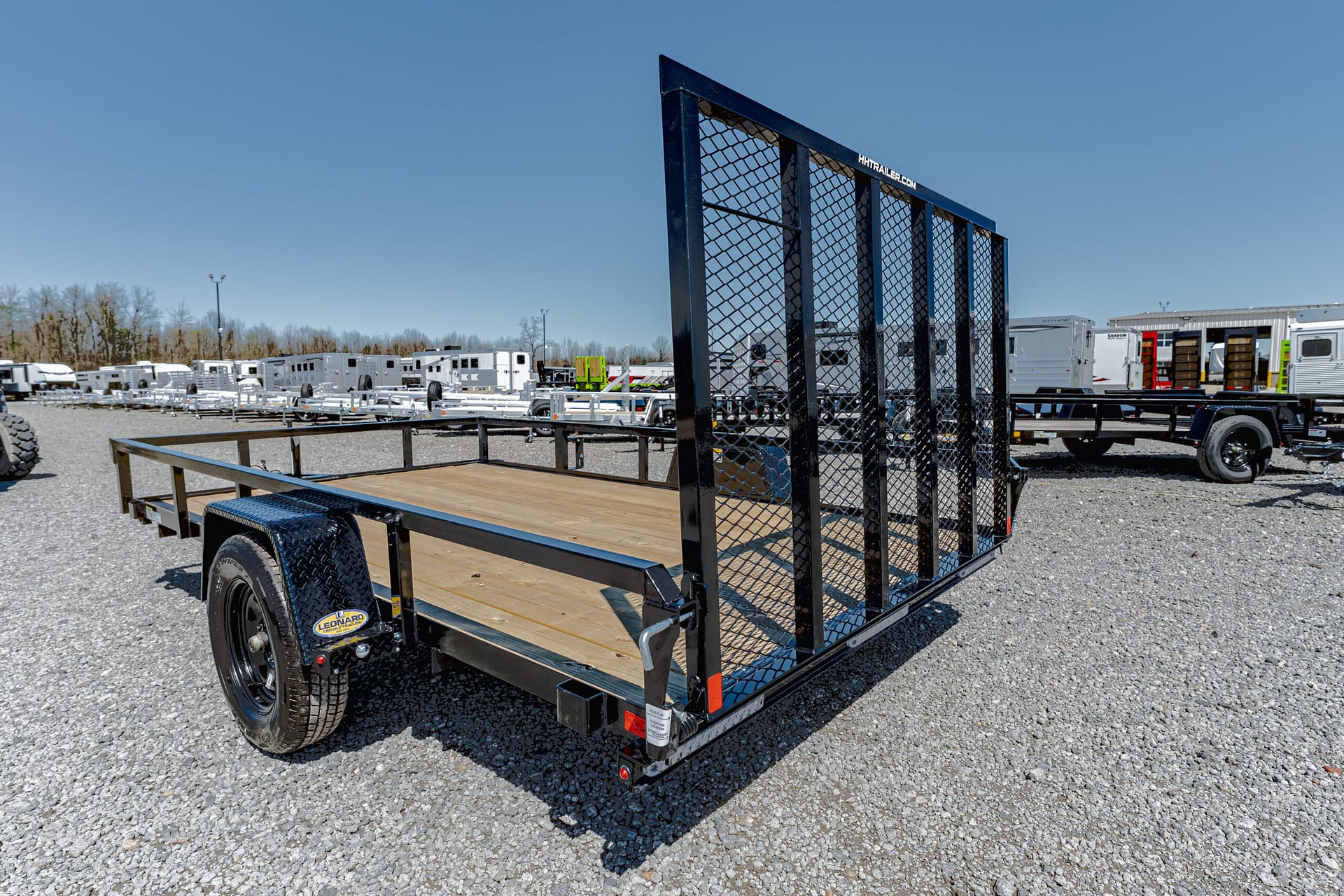 Flatbed utility trailer with a tall mesh ramp gate, wooden deck, and single axle, parked on gravel in a trailer lot.