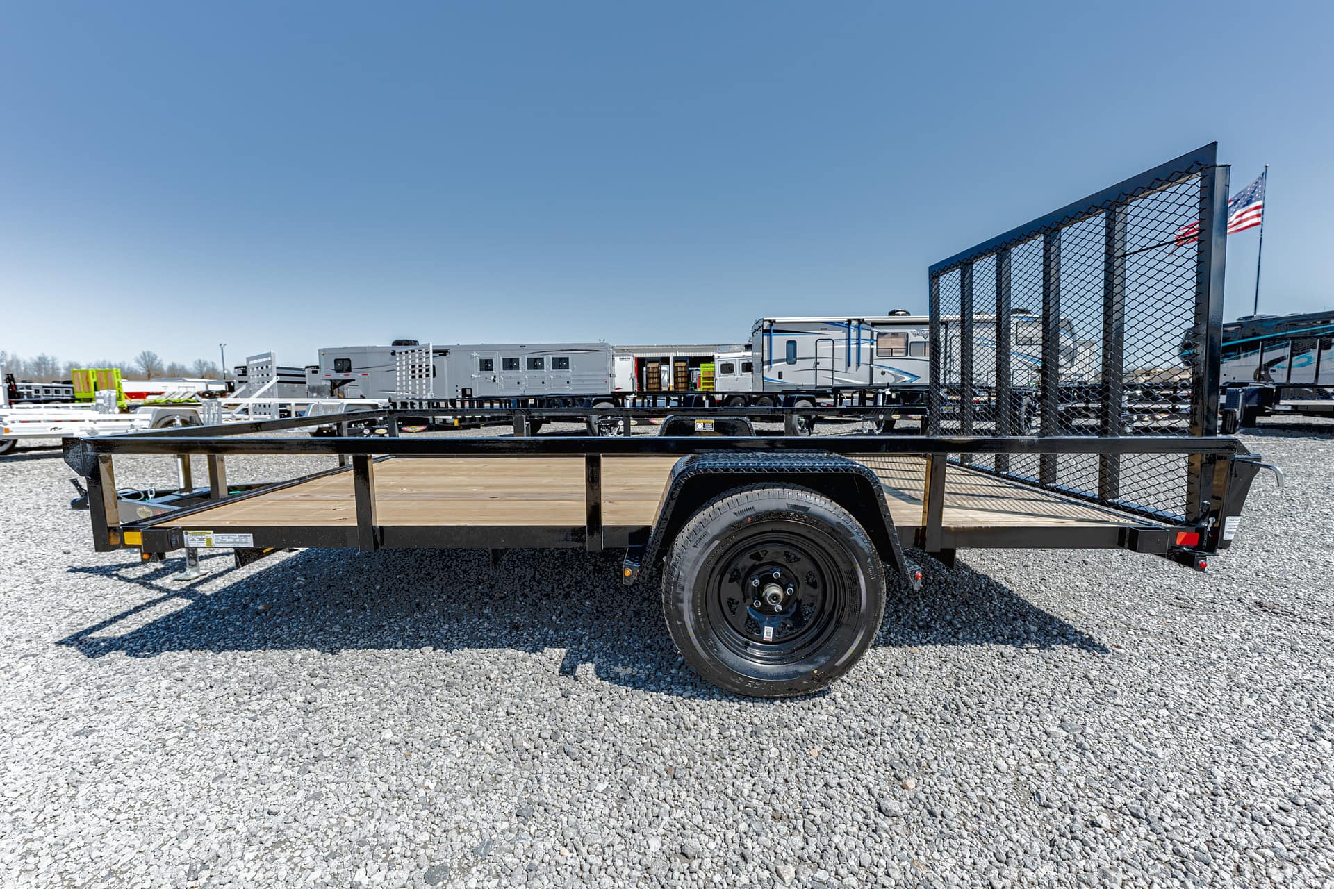 Flatbed utility trailer with wooden deck and mesh ramp gate parked on gravel, with other trailers in the background and an American flag nearby.