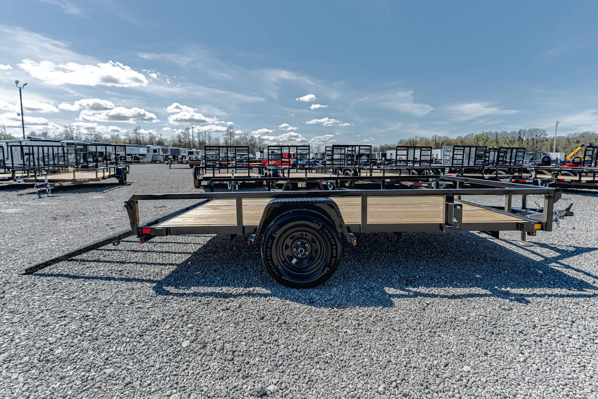 A flatbed utility trailer with a wooden bed and black frame parked in a gravel lot among many other trailers under a blue sky.