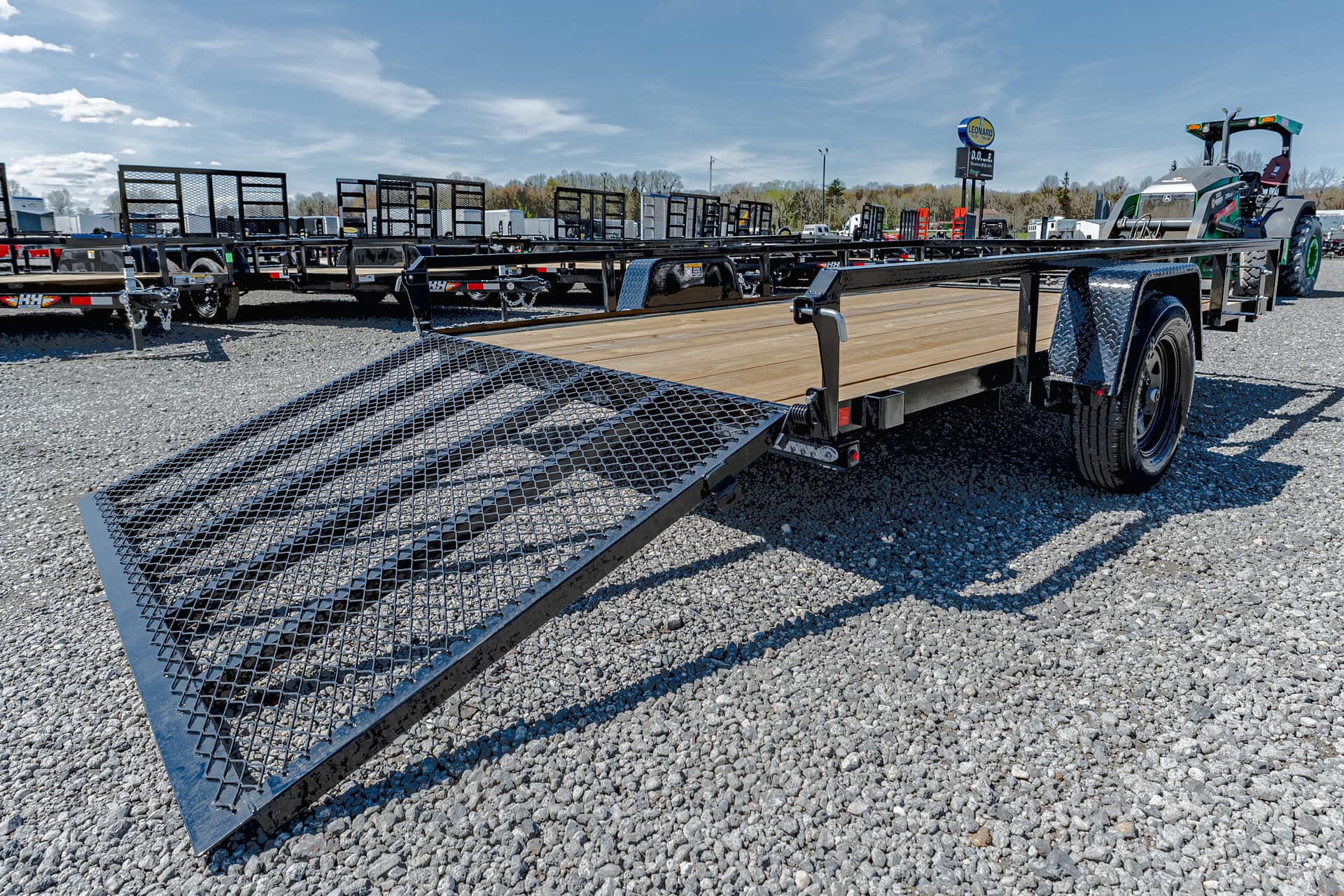 Flatbed trailer with a grated, fold-down loading ramp extended on a gravel lot, ready for hauling equipment.
