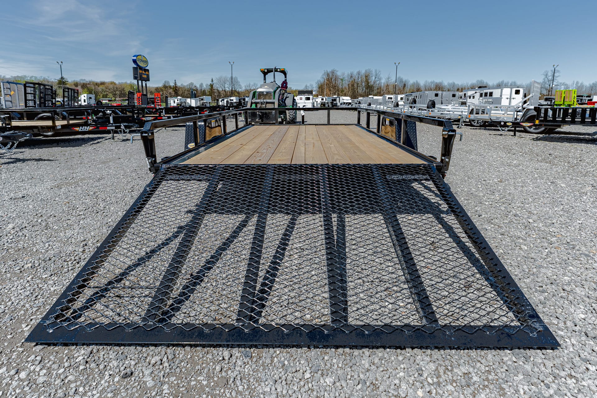 Black metal utility trailer with a wooden deck and diamond-mesh ramp, parked in a gravel lot with other trailers in the background.