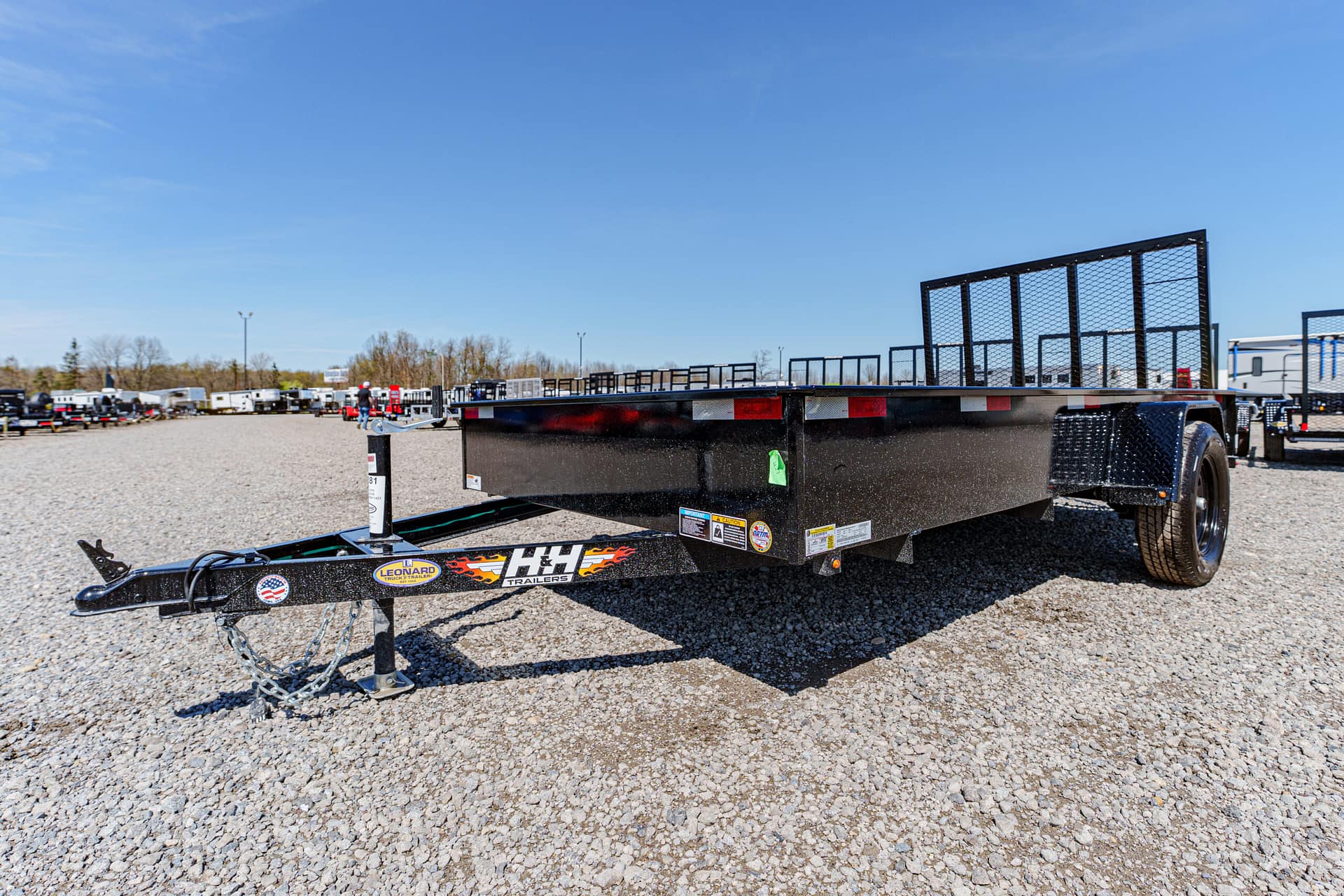 Black flatbed trailer with guard rails sits on a gravel lot, blue sky above and other trailers in the background.