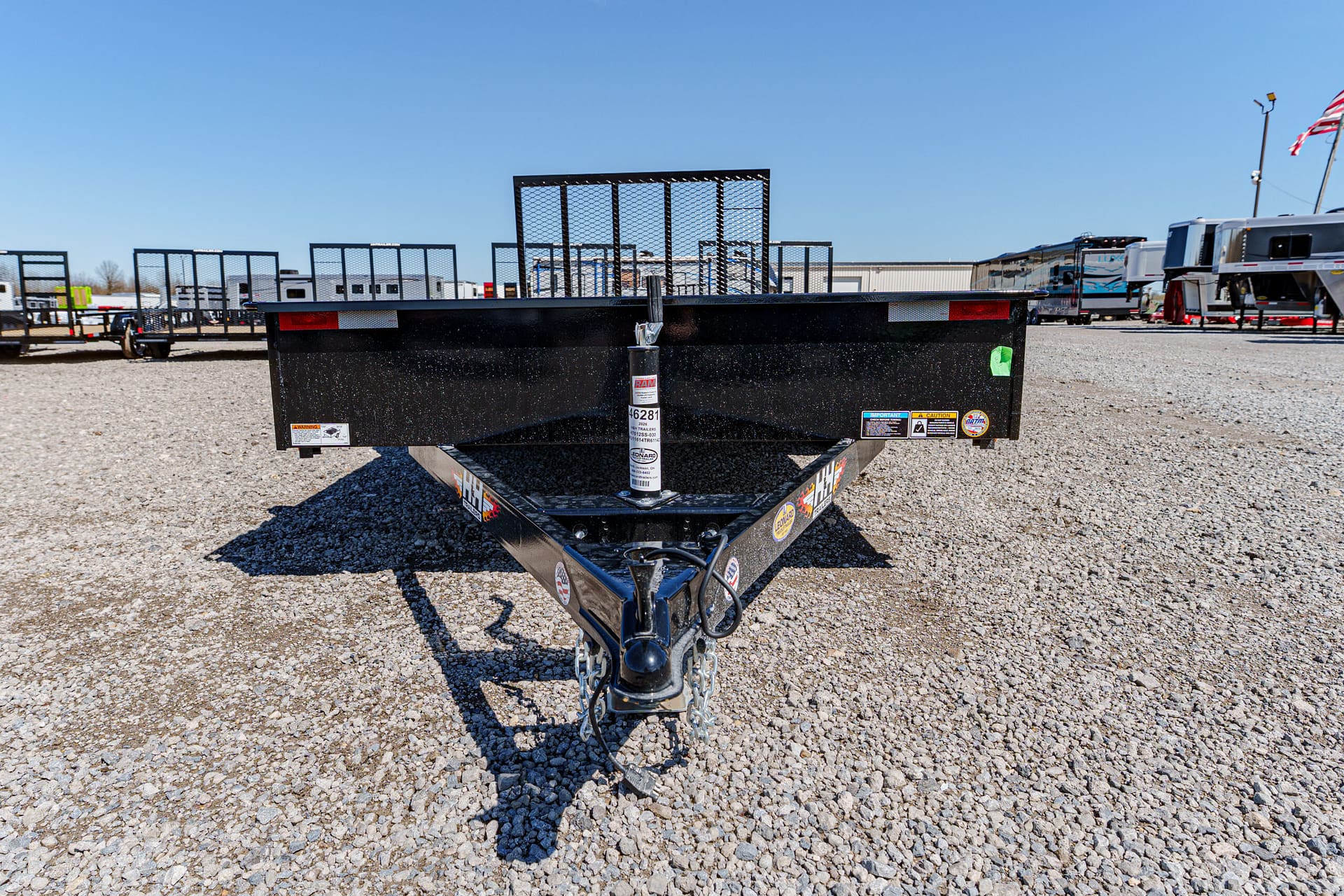 Close-up of a black trailer hitch and safety chains on a gravel lot, with other trailers in the background.