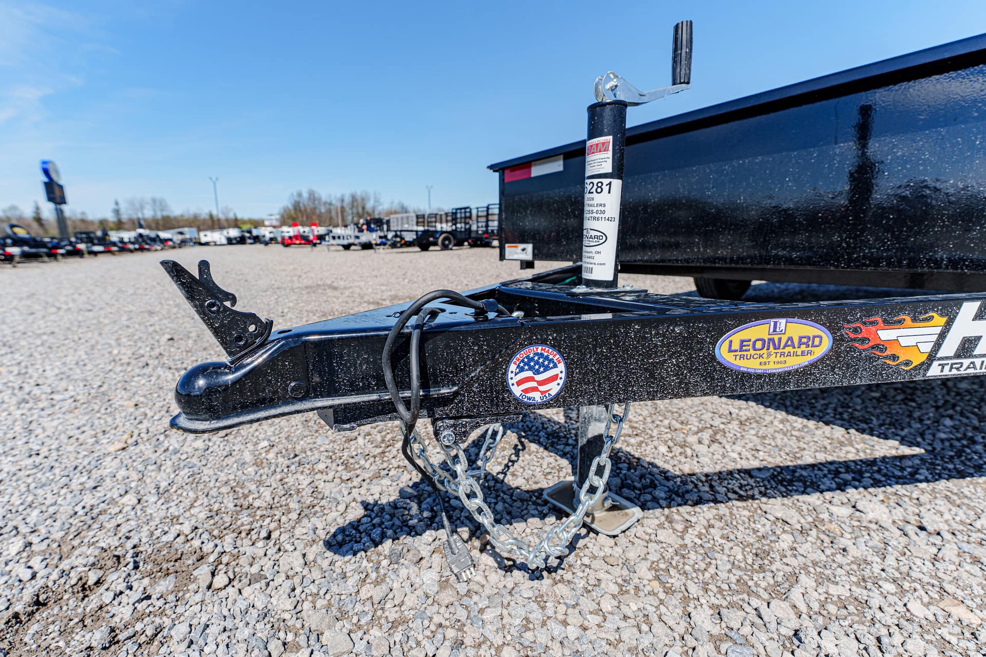 Close-up of a black trailer hitch with safety chains on a gravel lot; decals and a jack post are visible in the background.