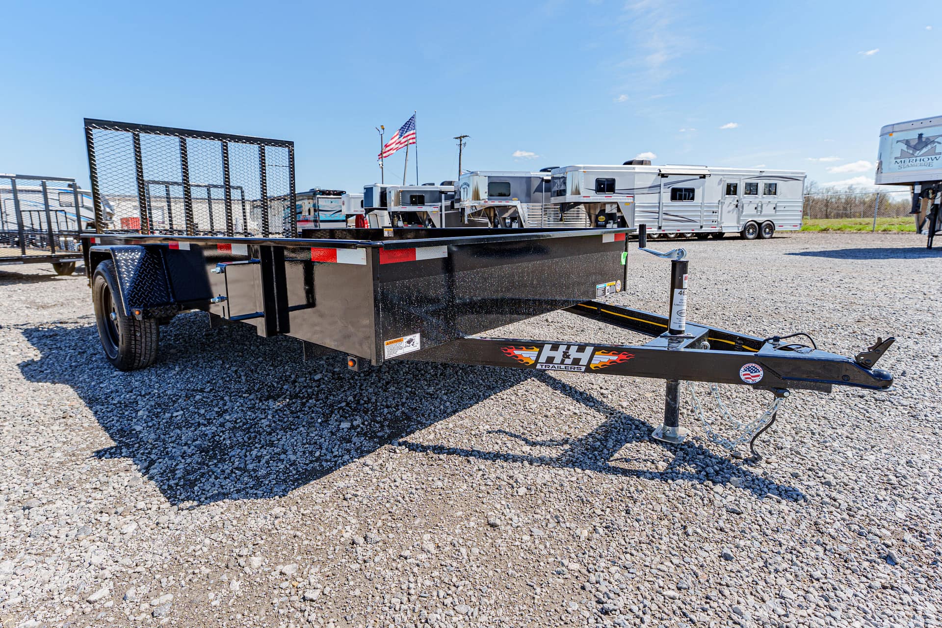 Black flatbed trailer with a mesh rear gate on a gravel lot, with RV trailers in the background and a blue sky above.